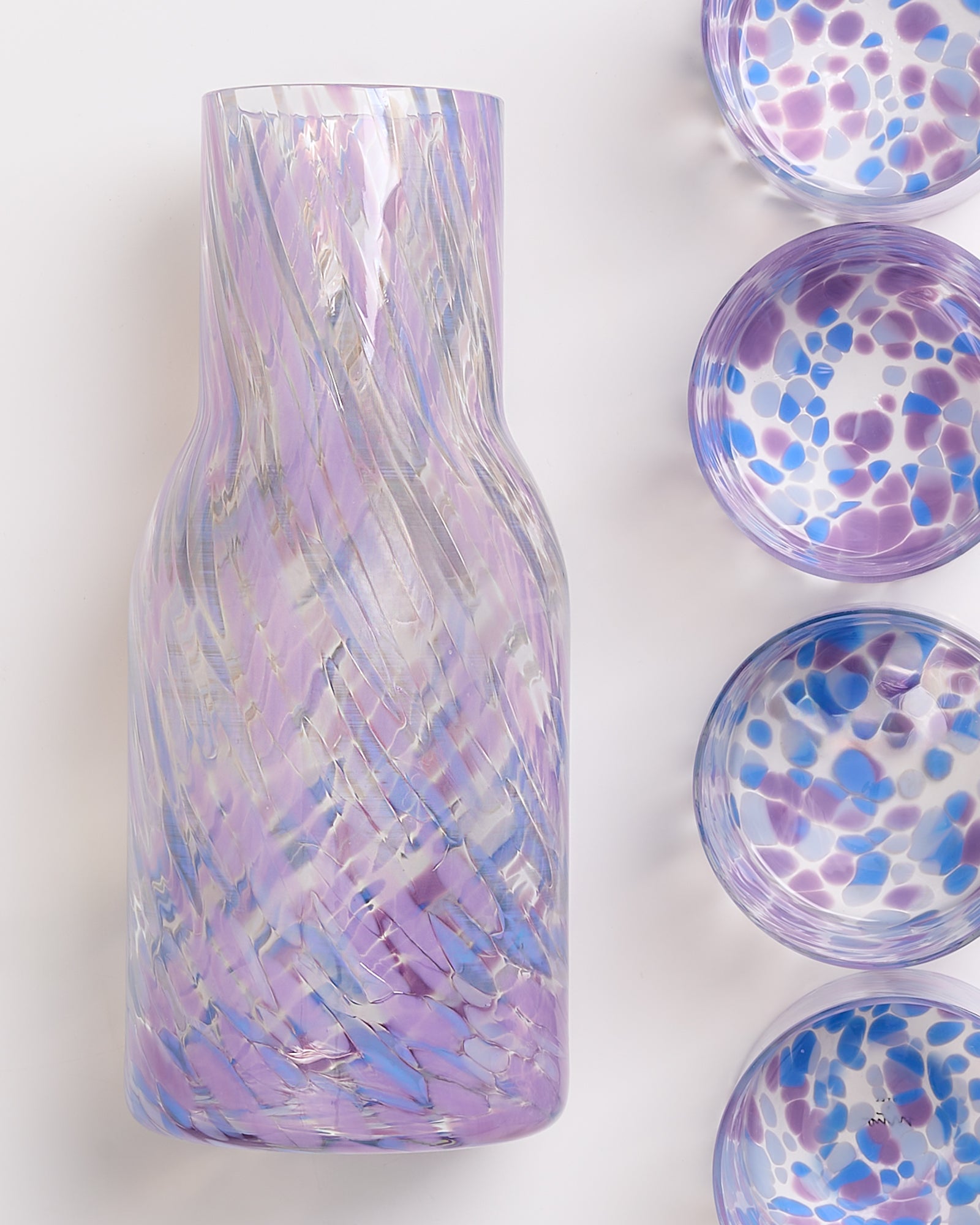 Violette Karaffe mit wirbelndem Muster und vier passende Schalen in lila und blau auf weißem Hintergrund.
