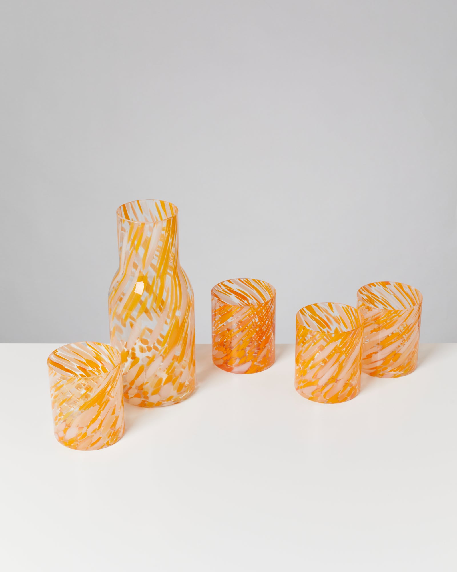 Fünf orange-weiß gemusterte Trinkgläser stehen auf einem weißen Tisch vor grauem Hintergrund.