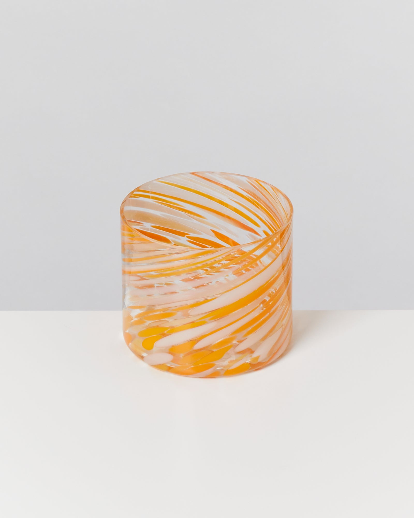 Zylinder aus Glas mit orange-weißen Wirbelmustern auf einer weißen Oberfläche, vor grauem Hintergrund.