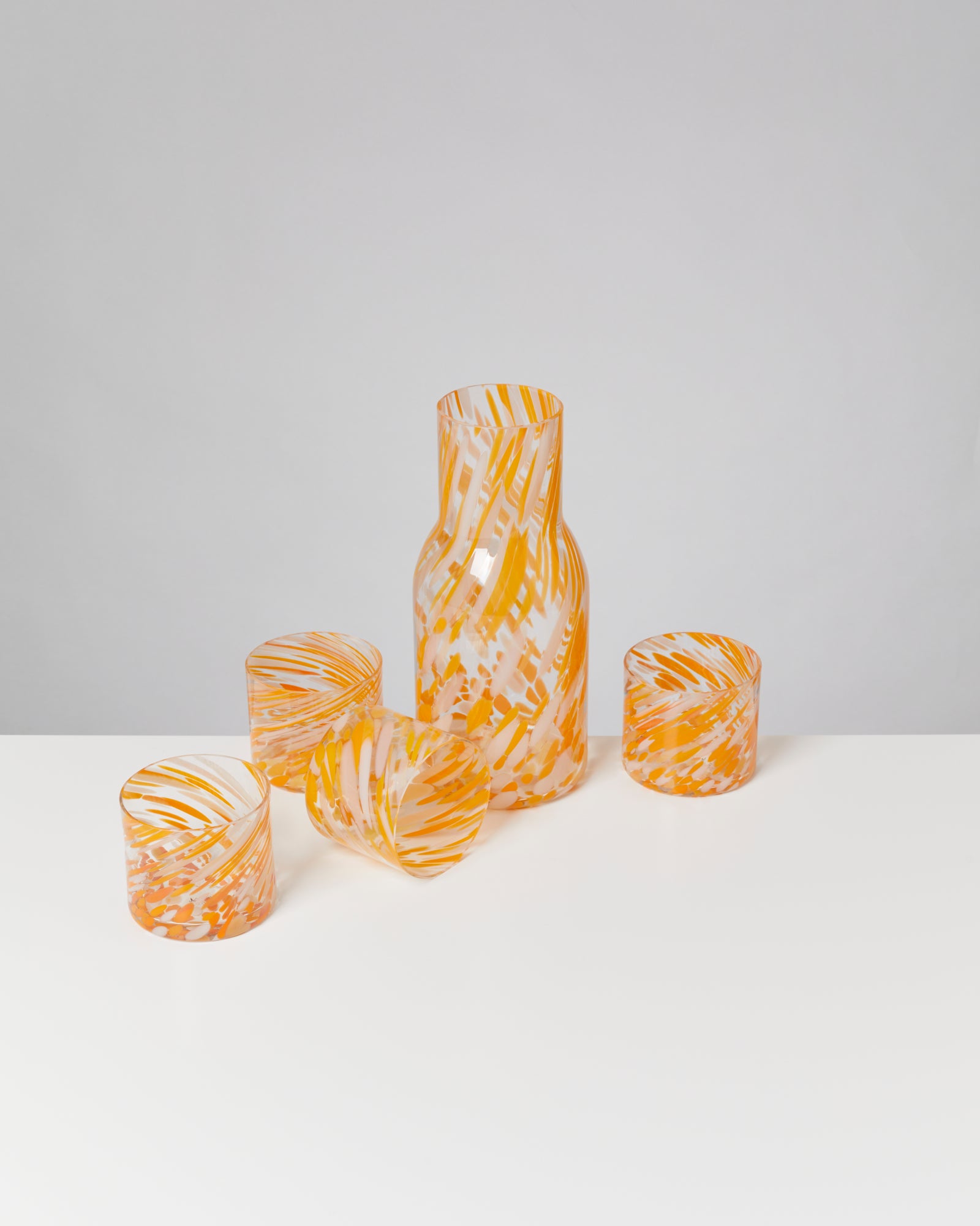 Karaffe und Gläser aus Glas mit orange-weißen Mustern stehen auf einem weißen Tisch vor grauem Hintergrund.