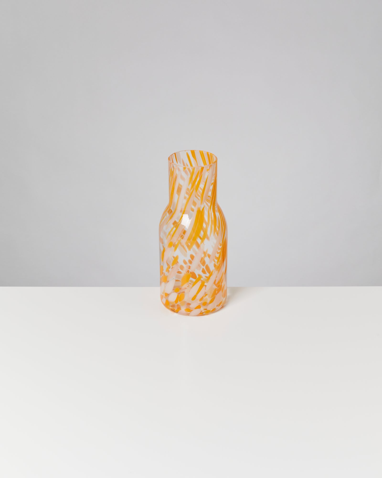 Bunte, orange-weiße Glasvase mit abstrakten Mustern steht auf einem weißen Tisch vor grauem Hintergrund.