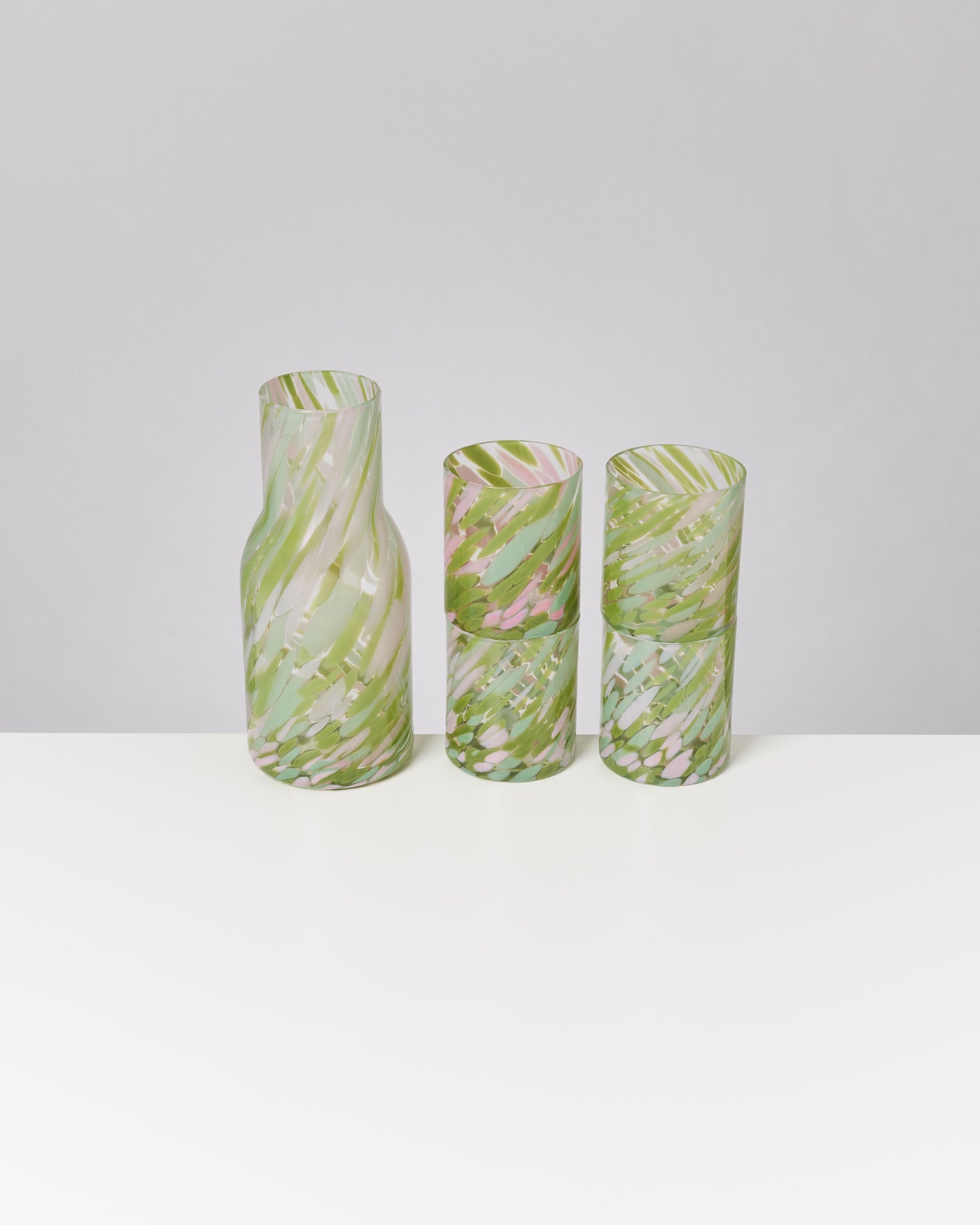 Drei gemusterte Glasgefäße in Grün- und Rosatönen stehen auf einem weißen Tisch vor grauem Hintergrund.
