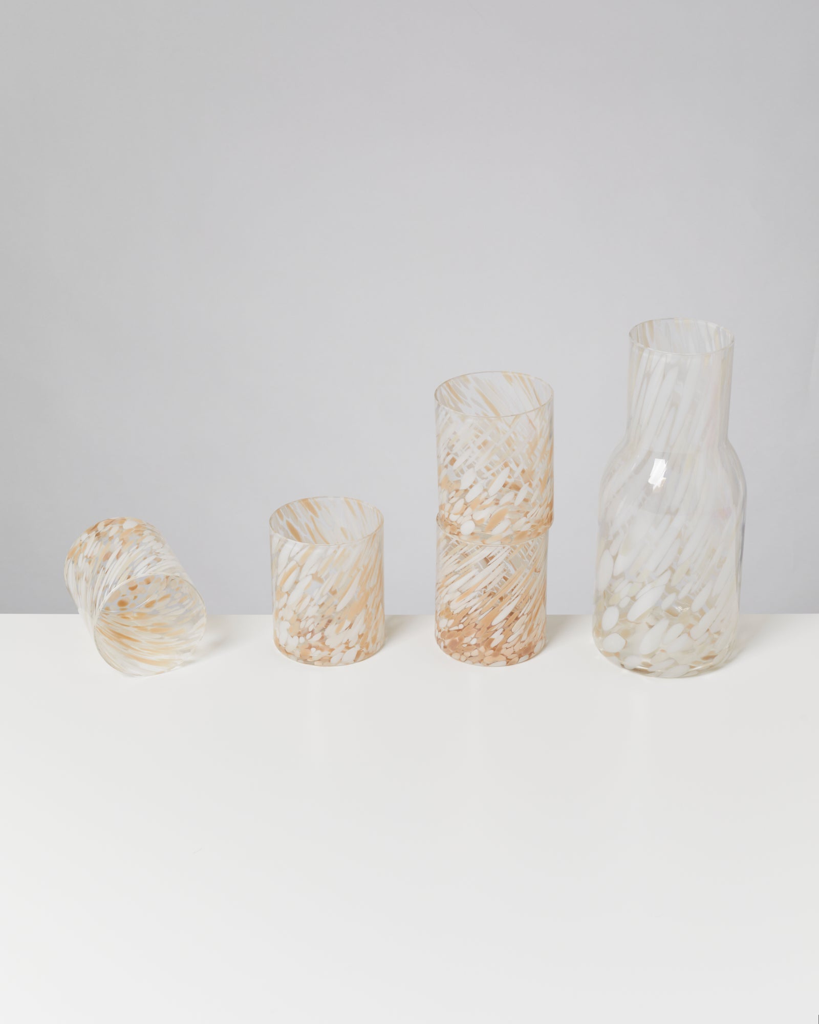 Vier Glasbecher und eine Karaffe mit weiß-goldenem Muster stehen auf einem weißen Tisch vor einem grauen Hintergrund.