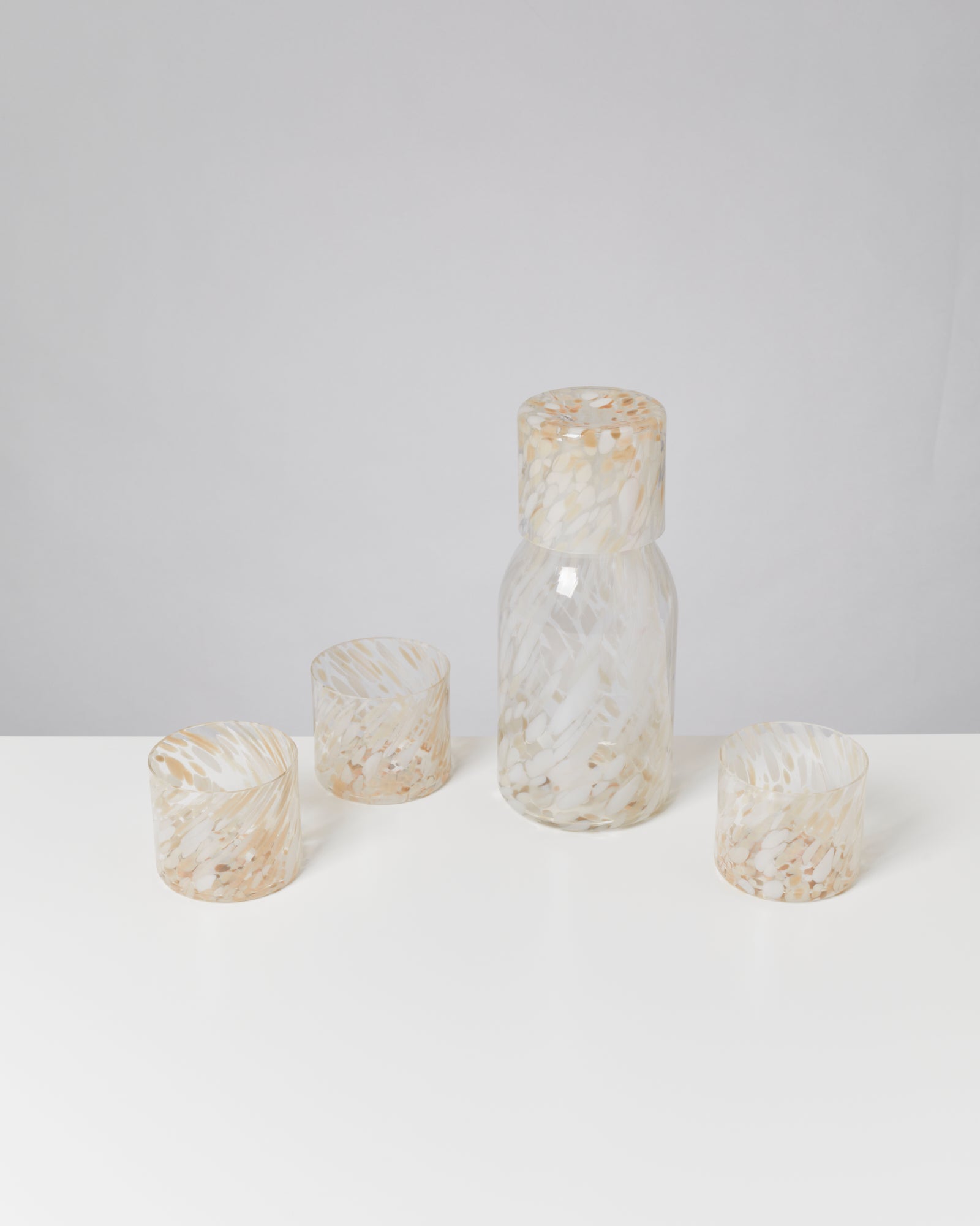 Glasflasche mit drei passenden Gläsern, alle mit beige-weißer Fleckenstruktur, auf weißem Hintergrund.