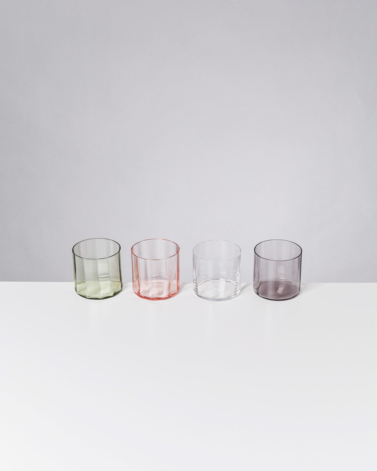 Vier bunte Trinkgläser in Grün, Rot, Klar und Grau stehen auf einem weißen Tisch vor grauem Hintergrund.