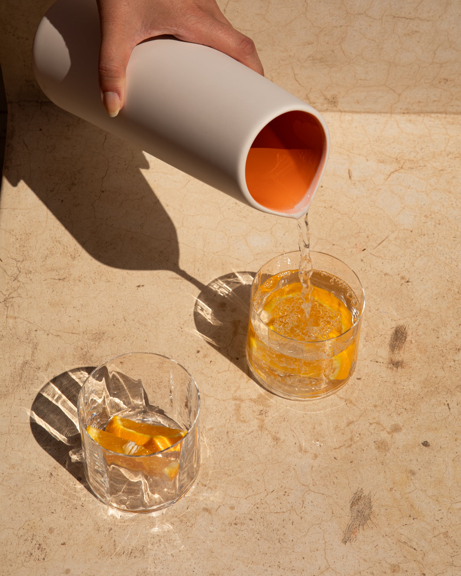 Hand gießt Wasser aus weißer Karaffe in zwei Gläser mit Orangenstücken auf einer hellen Oberfläche.