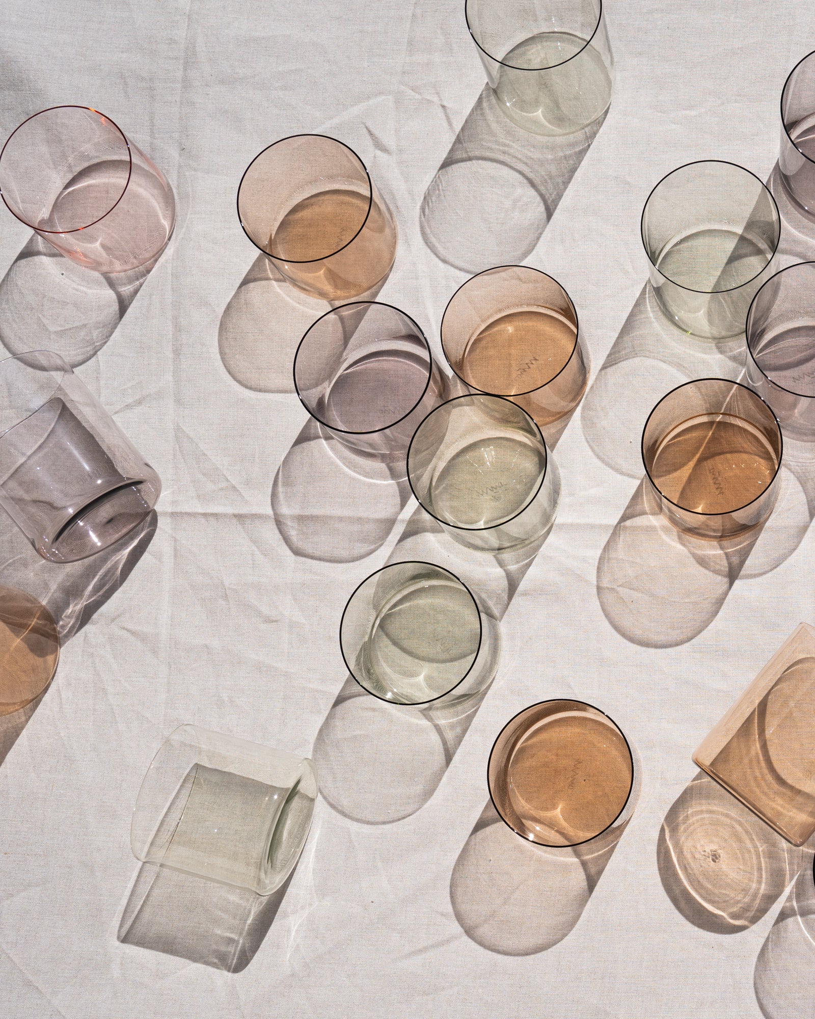 Mehrere transparente, bunte Gläser liegen verstreut auf einem hellen Stofftuch, mit Schattenwürfen sichtbar.