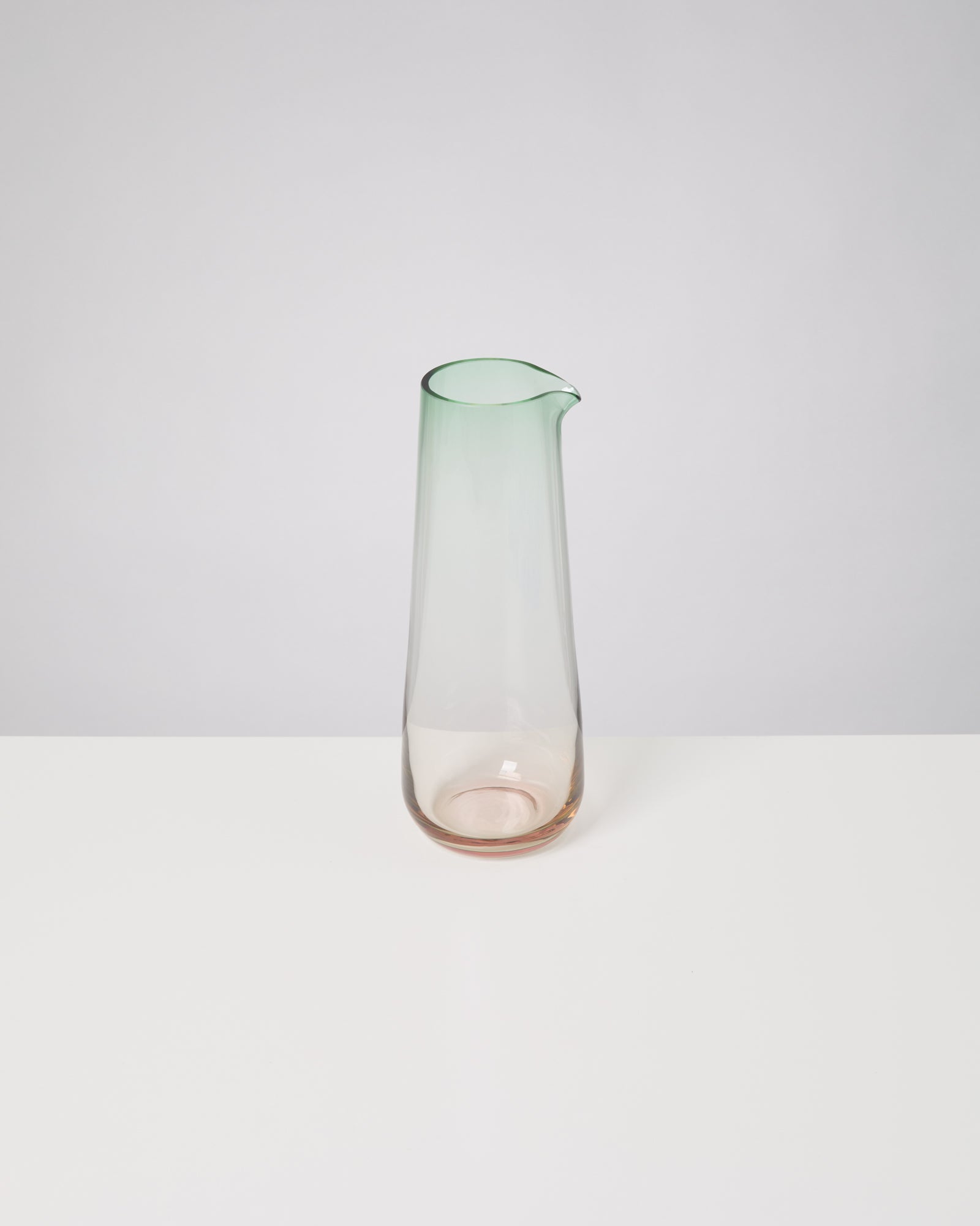 Ein transparenter Krug mit grünlichem oberen Rand steht auf einem weißen Tisch vor einem grauen Hintergrund.