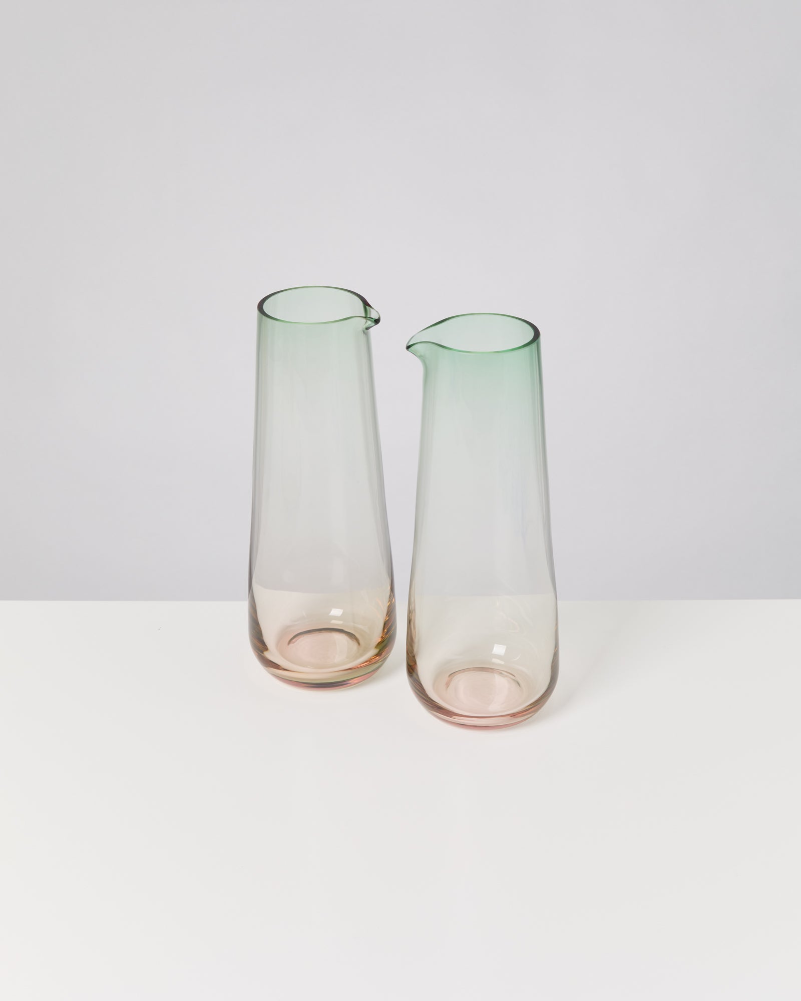 Zwei schlichte Glasgefäße mit grünem Rand und schmalem Hals stehen auf einer weißen Oberfläche.