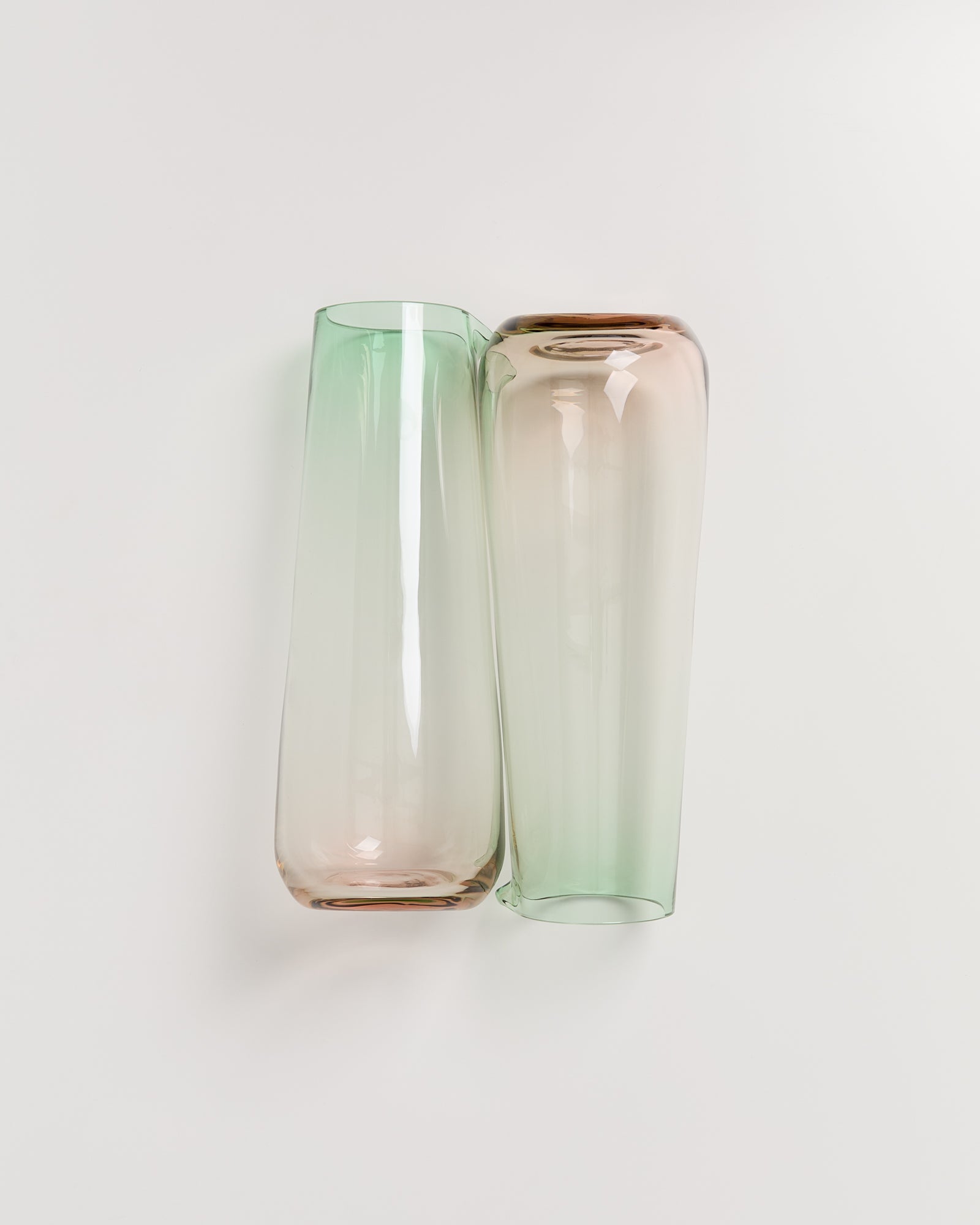 Zwei schlanke Glasvasen in Pastelltönen, eine grün und die andere rosa, nebeneinander auf weißem Hintergrund.