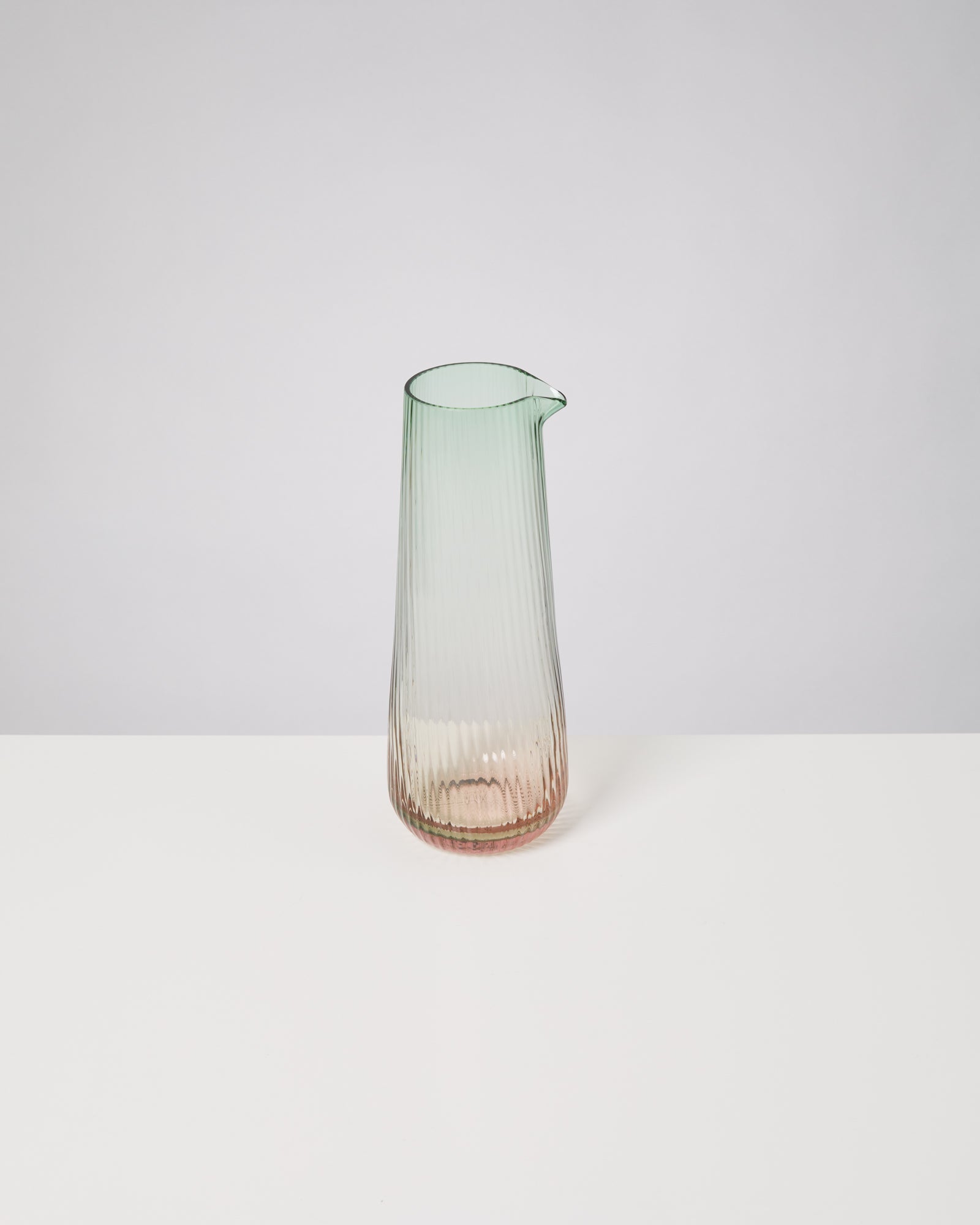 Durchsichtige Glaskaraffe mit Farbverlauf von Grün nach Rosa, auf einem weißen Tisch vor weißem Hintergrund.