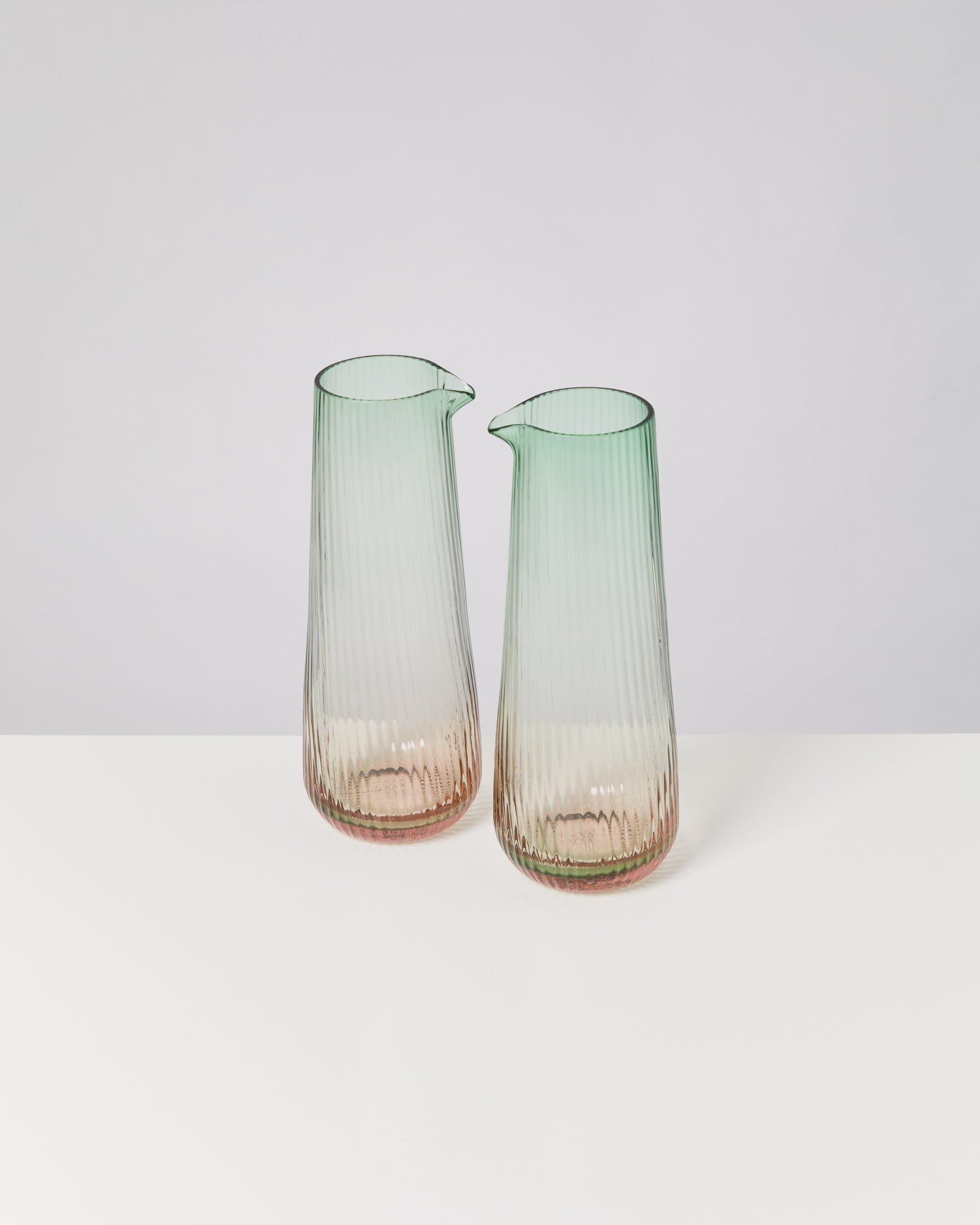 Zwei gerippte Glasvasen mit grünlichem Farbverlauf auf einem weißen Tisch vor grauem Hintergrund.