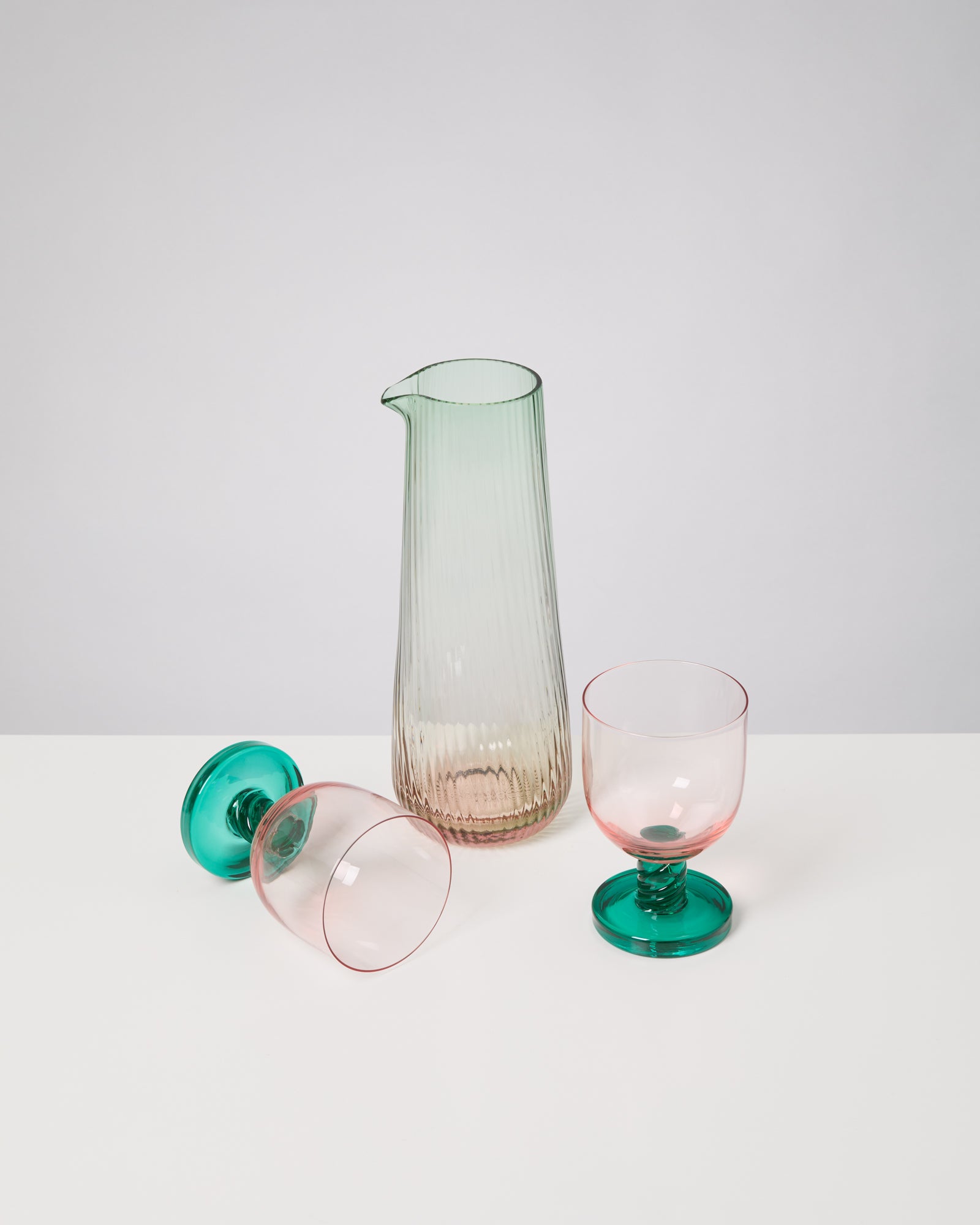 Grüne Glaskaraffe und zwei rosafarbene Gläser mit grünen Stielen, eines liegend, auf weißem Hintergrund.