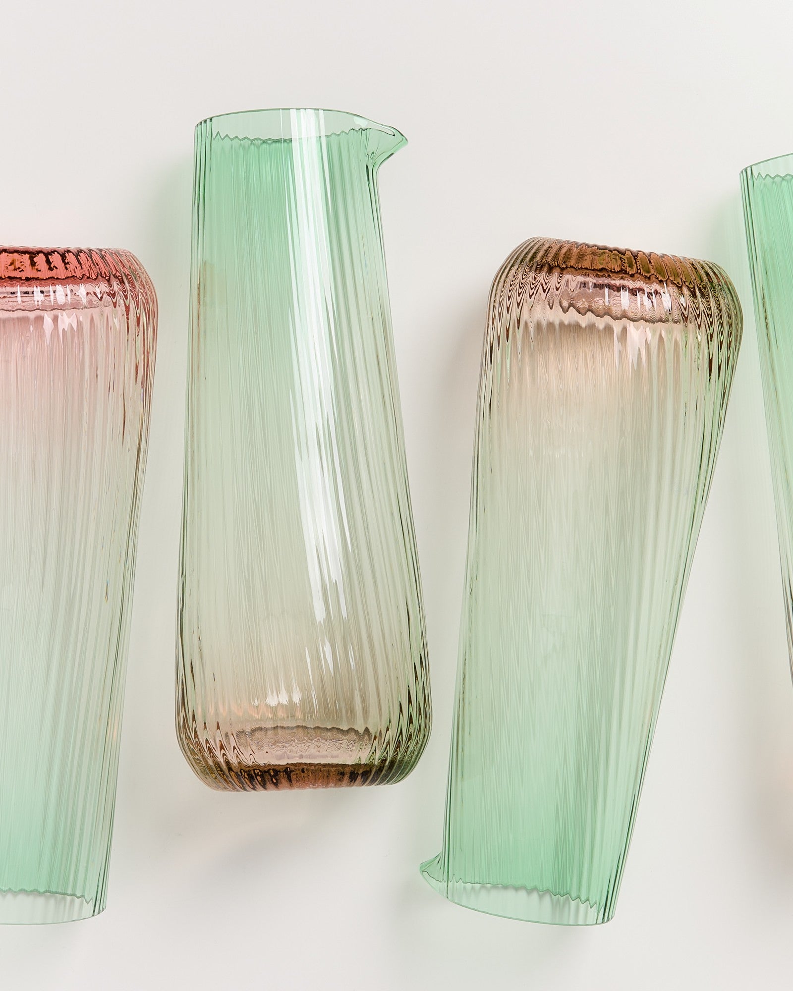 Vier schräg stehende Glaskaraffen in Rosé und Grün mit Rillenstruktur vor weißem Hintergrund.
