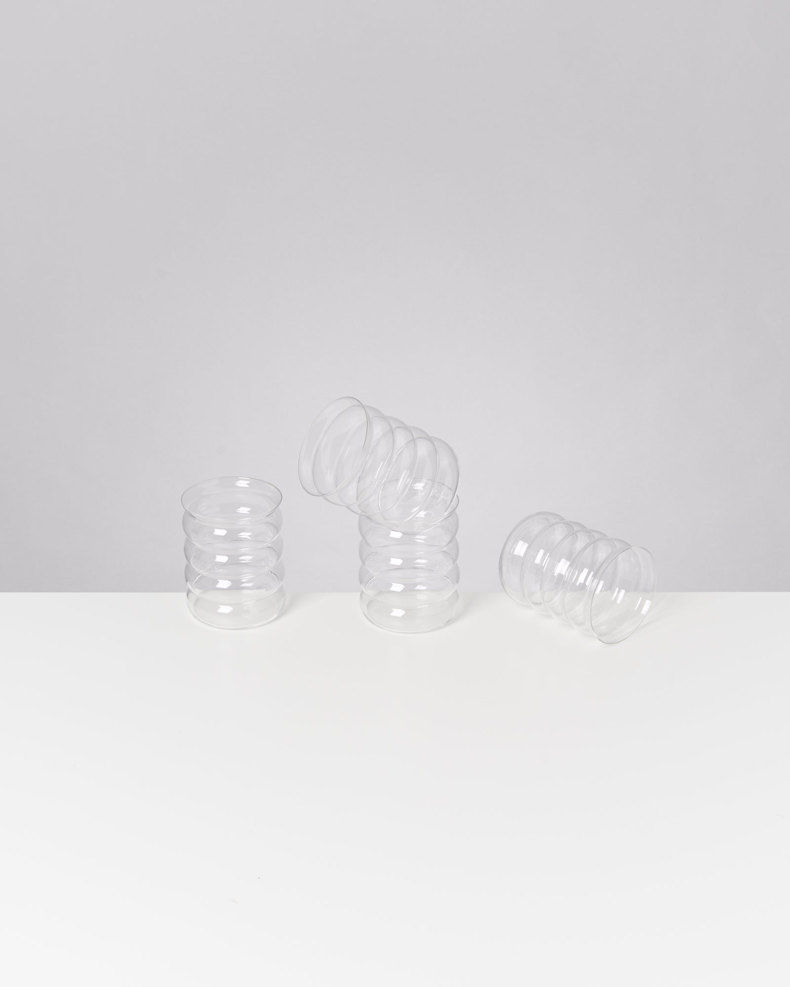 Drei durchsichtige, spiralförmige Gläser auf einem weißen Tisch, eines davon ist umgekippt.