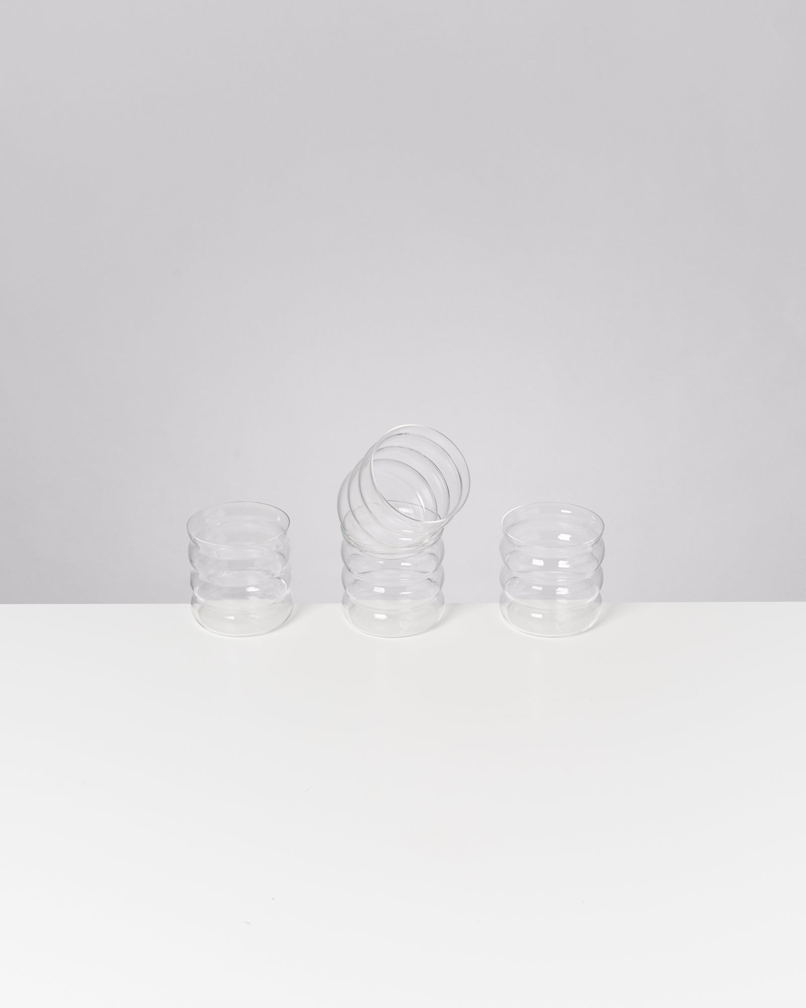 Drei klare Gläser mit spiralartigem Design stehen auf einem weißen Tisch, wobei eines leicht zur Seite geneigt ist.