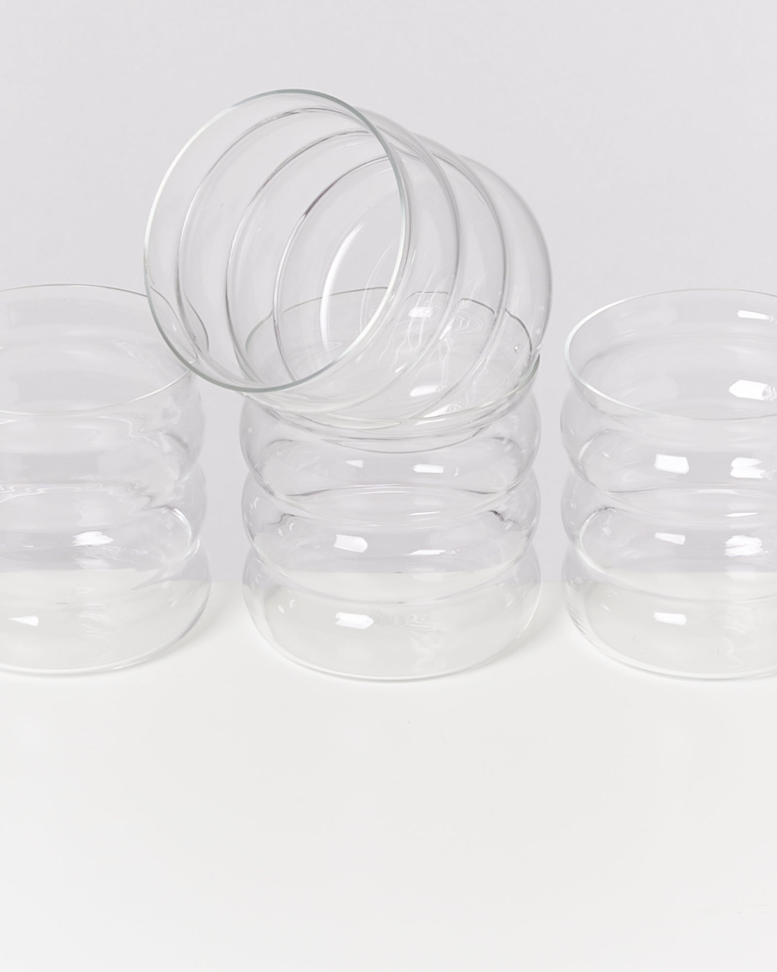 Vier gläserne Becher, einer liegt schräg auf drei anderen, auf weißem Hintergrund.