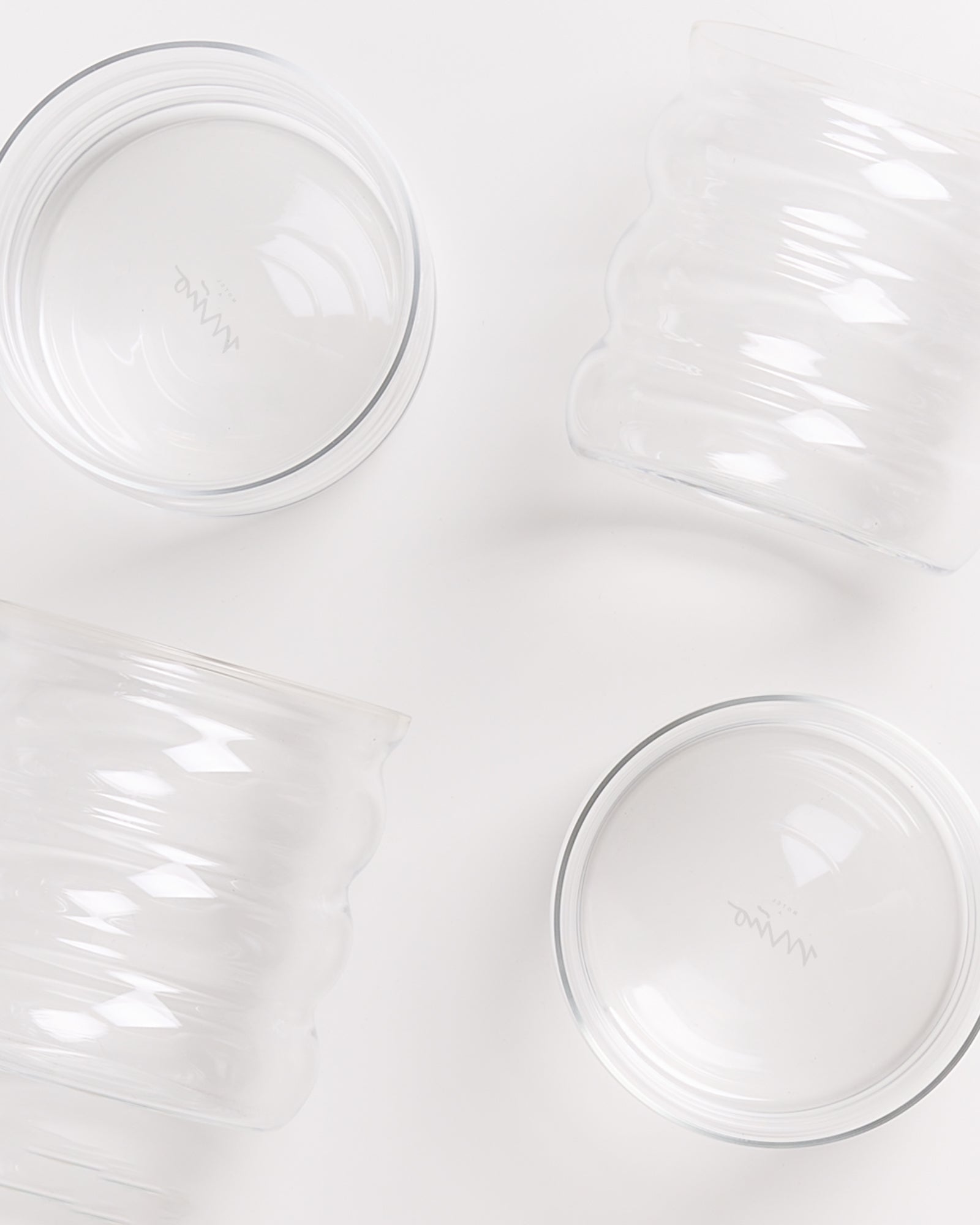 Vier transparente Glasbehälter auf einer weißen Oberfläche, zwei rund und zwei gewellt geformt.