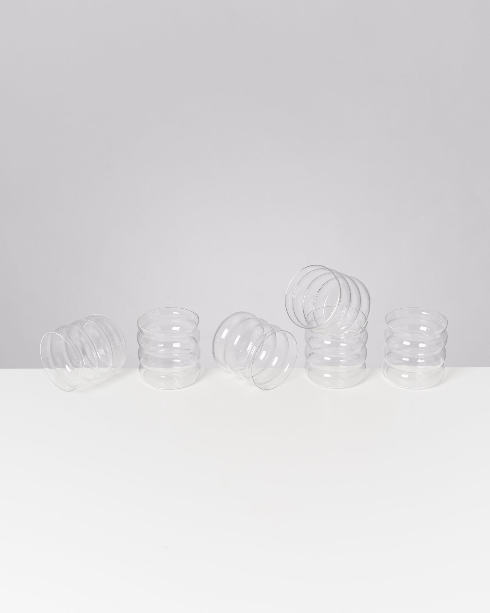 Fünf durchsichtige, gewellte Gläser stehen und liegen aufrecht auf einem weißen Tisch vor grauem Hintergrund.