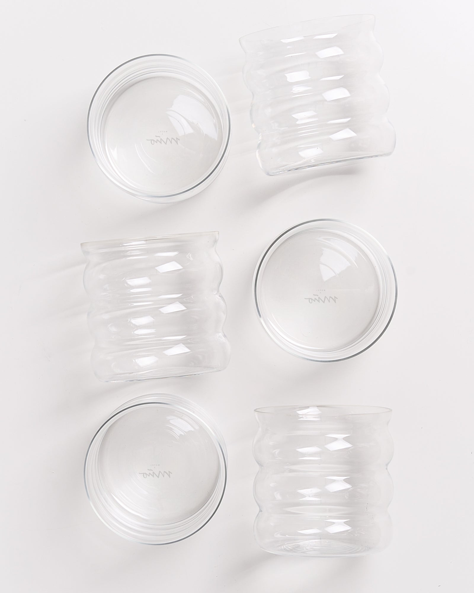 Sechs wellenförmige, durchsichtige Gläser auf einer weißen Unterlage, teilweise übereinander gestapelt.