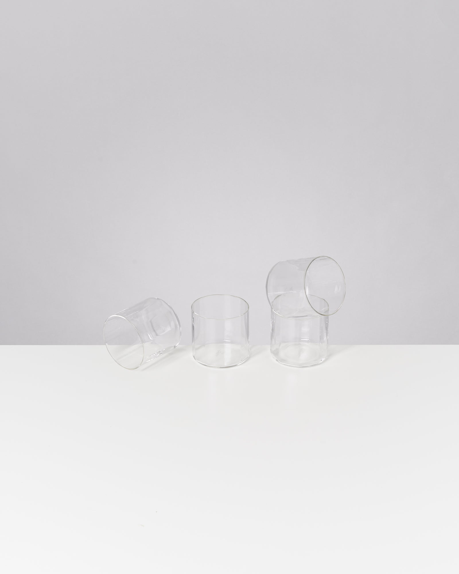 Vier leere Gläser stehen und liegen auf einer weißen Oberfläche vor einem grauen Hintergrund.