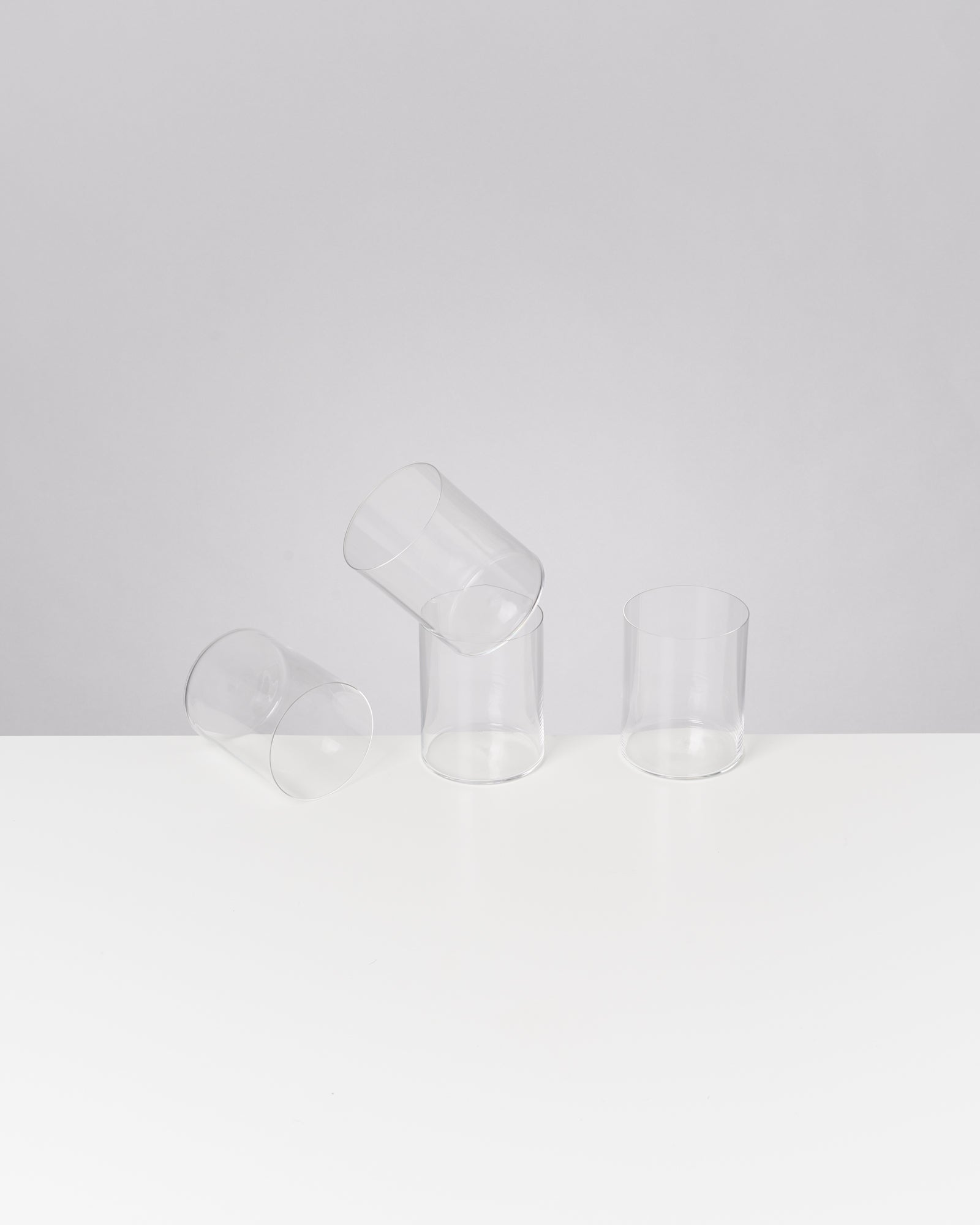 Drei durchsichtige Gläser auf weißem Tisch, zwei stehen, eines liegt schräg.