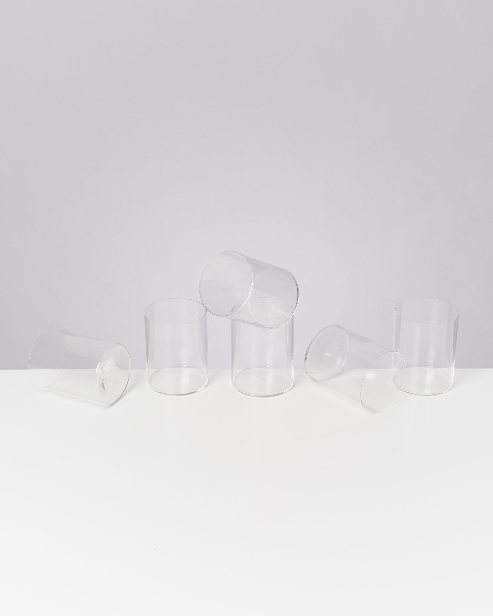 Fünf klare, zylindrische Gläser liegen und stehen auf einer weißen Oberfläche vor grauem Hintergrund.