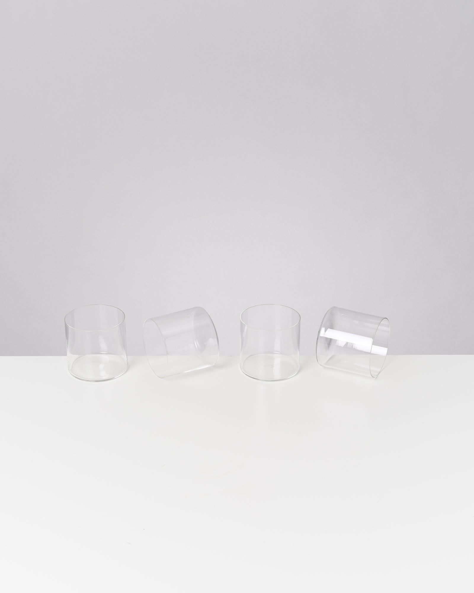 Vier transparente Gläser stehen in einer Reihe auf einem weißen Tisch, zwei davon liegen seitlich.