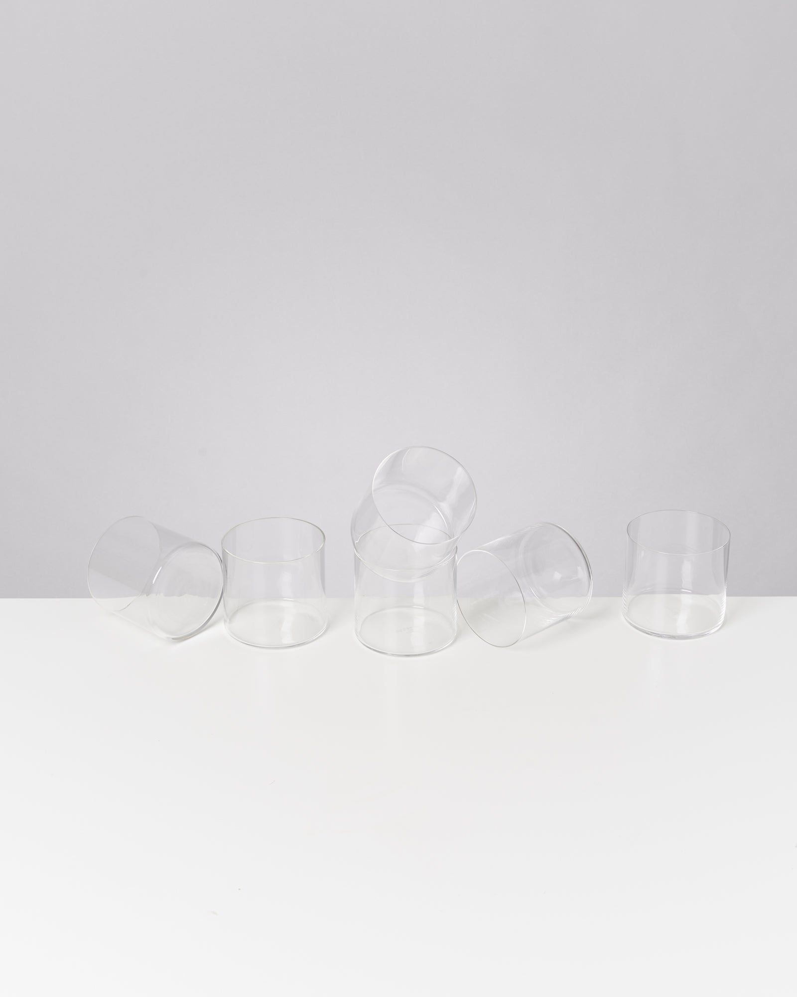 Sechs umgekippte, transparente Gläser auf einem weißen Tisch vor grauem Hintergrund.