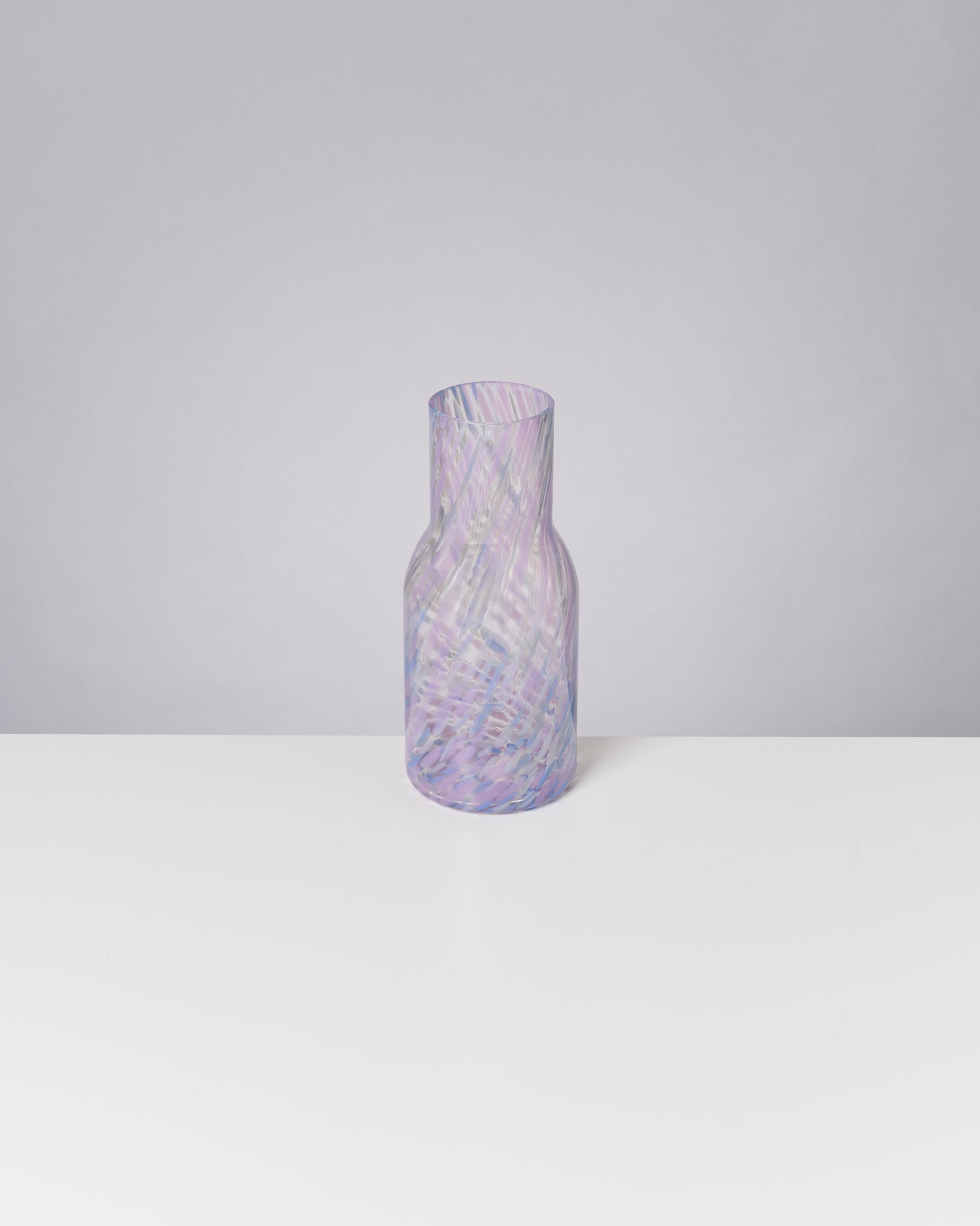 Ein gläserner, pastellfarbener Krug mit spiralförmigem Muster steht auf einem weißen Tisch vor hellgrauem Hintergrund.