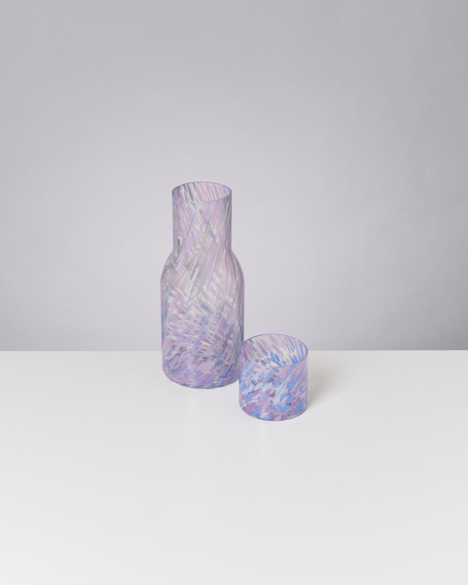 Eine transparente Karaffe mit passendem Glas, beide mit einem lila und bläulichen Muster, stehen auf einem weißen Tisch.