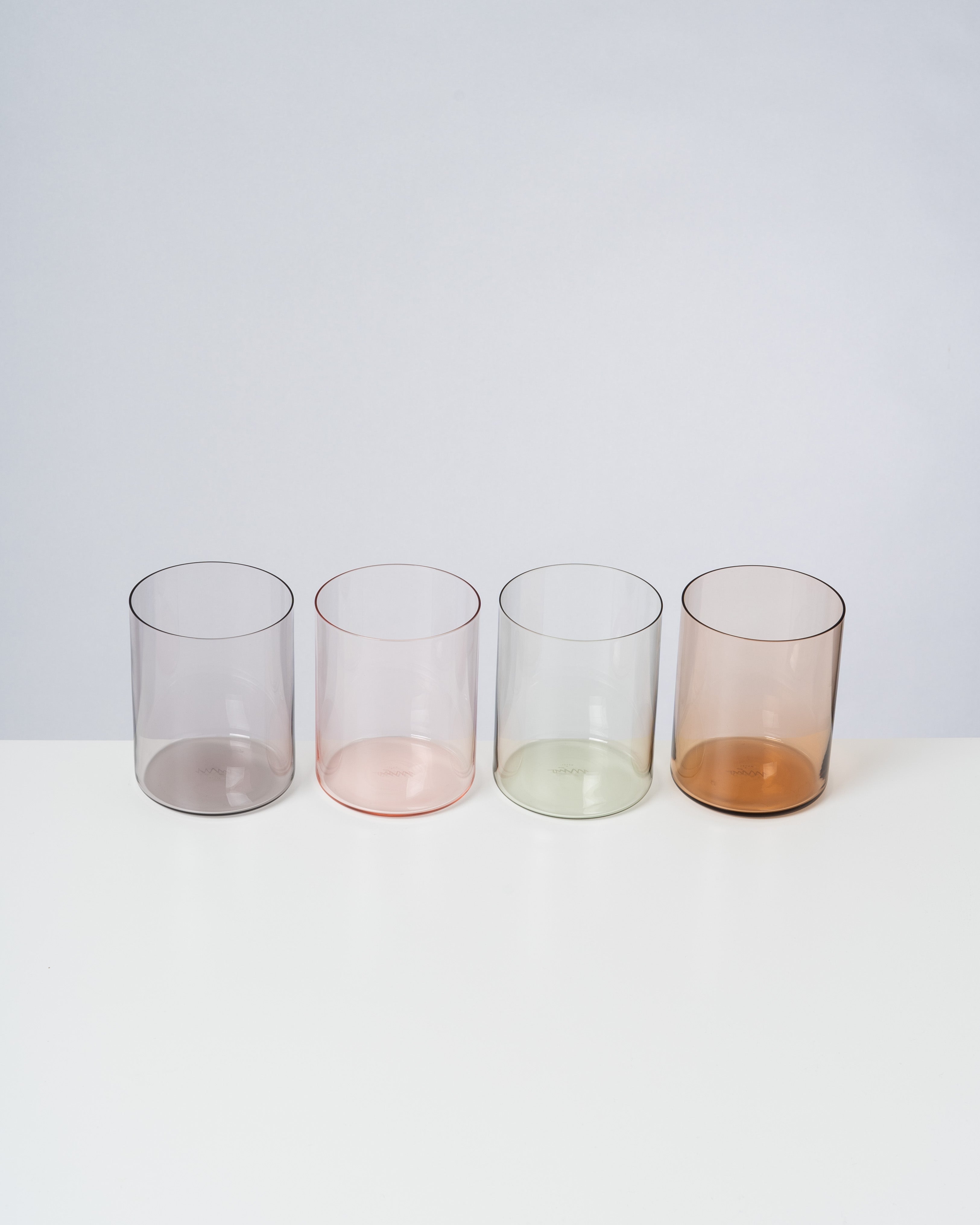 Vier farbige Glasbecher in Lila, Rosa, Grün und Braun stehen nebeneinander auf einem weißen Hintergrund.
