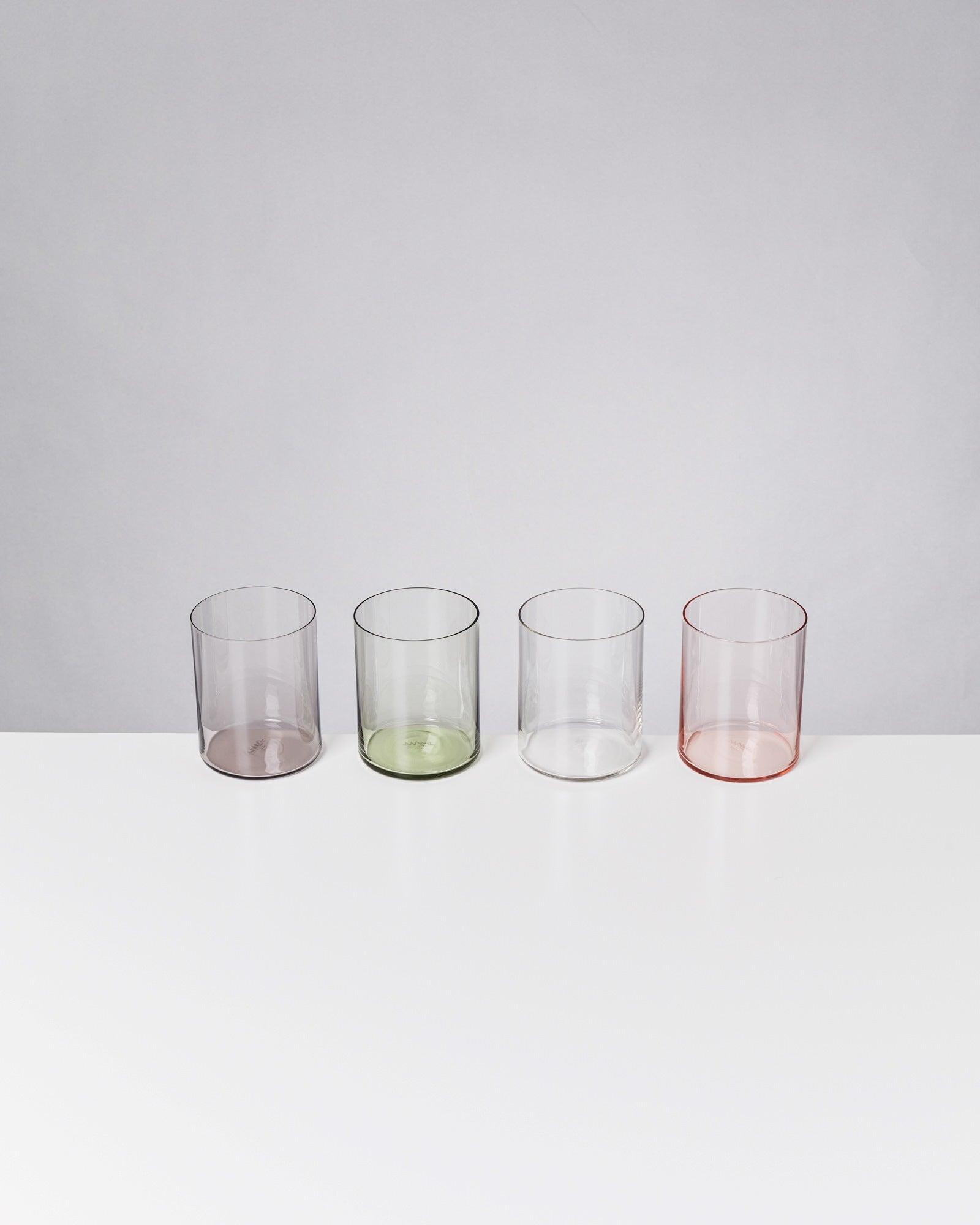 Vier transparente, farbige Gläser in Lila, Grün, Klar und Rosa auf einem weißen Tisch vor grauem Hintergrund.