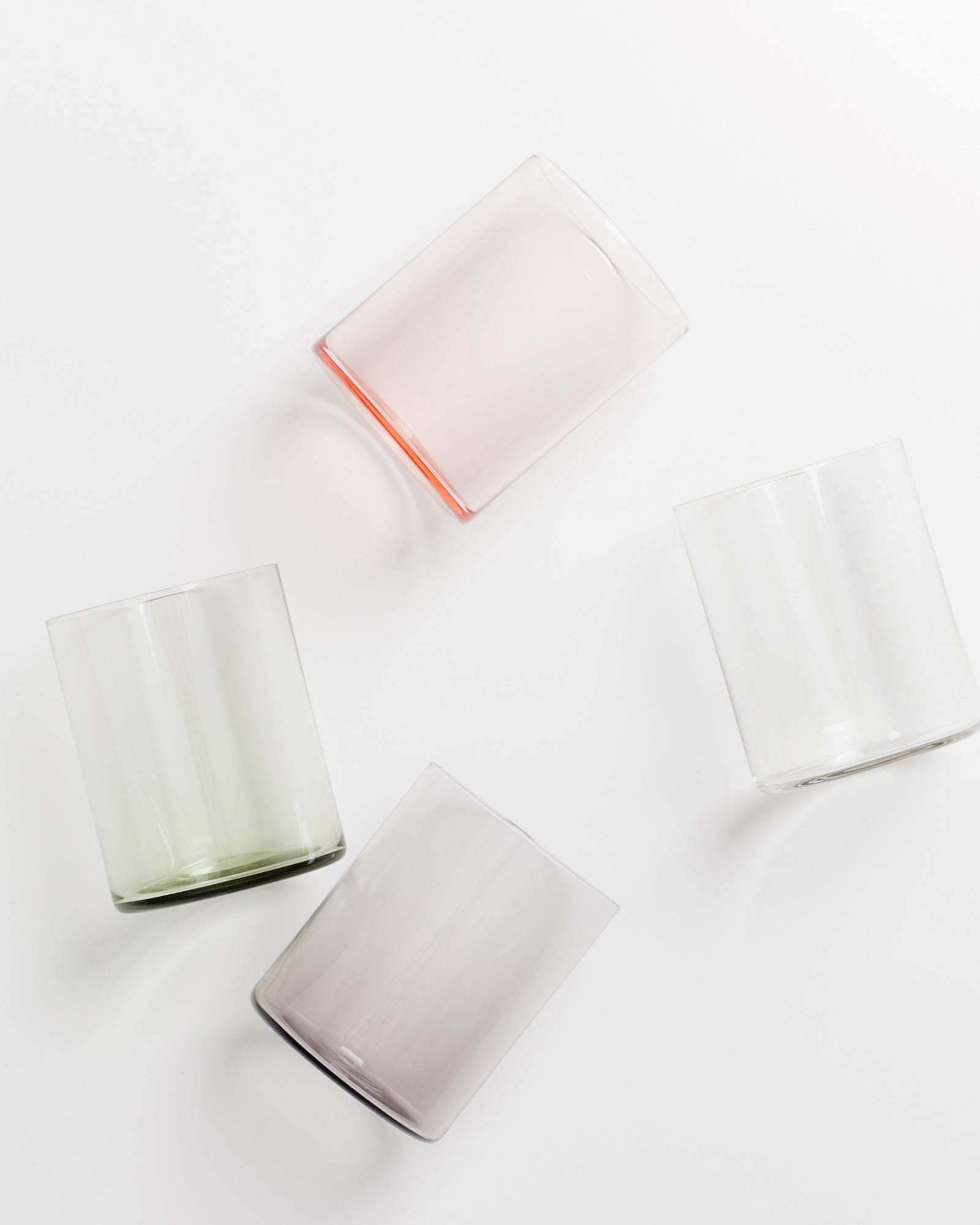 Vier bunte Trinkgläser liegen schräg verteilt auf einer weißen Oberfläche.