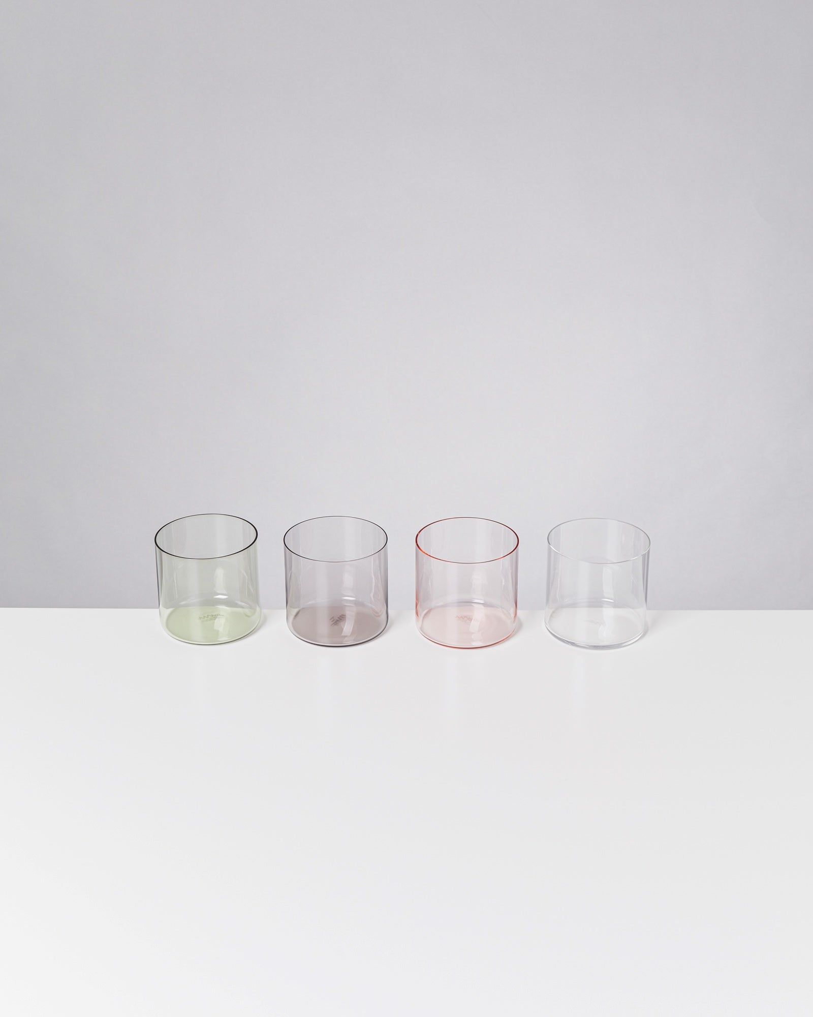 Vier farbige, zylindrische Gläser (grün, lila, rosa, durchsichtig) stehen auf einer weißen Oberfläche vor grauem Hintergrund.