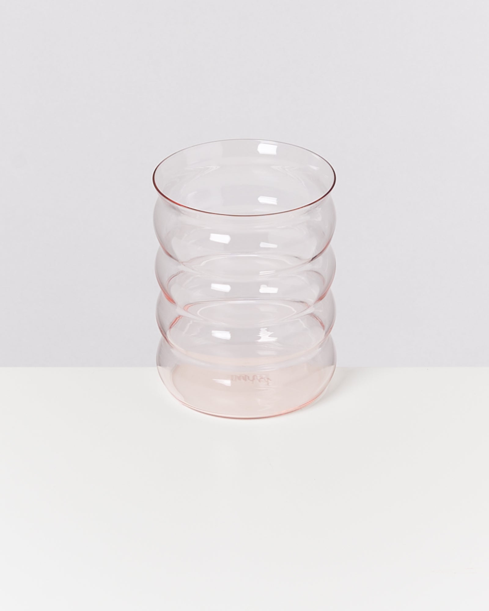 Ein transparentes Glas mit wellenförmigem Design steht auf einer weißen Oberfläche vor einem grauen Hintergrund.