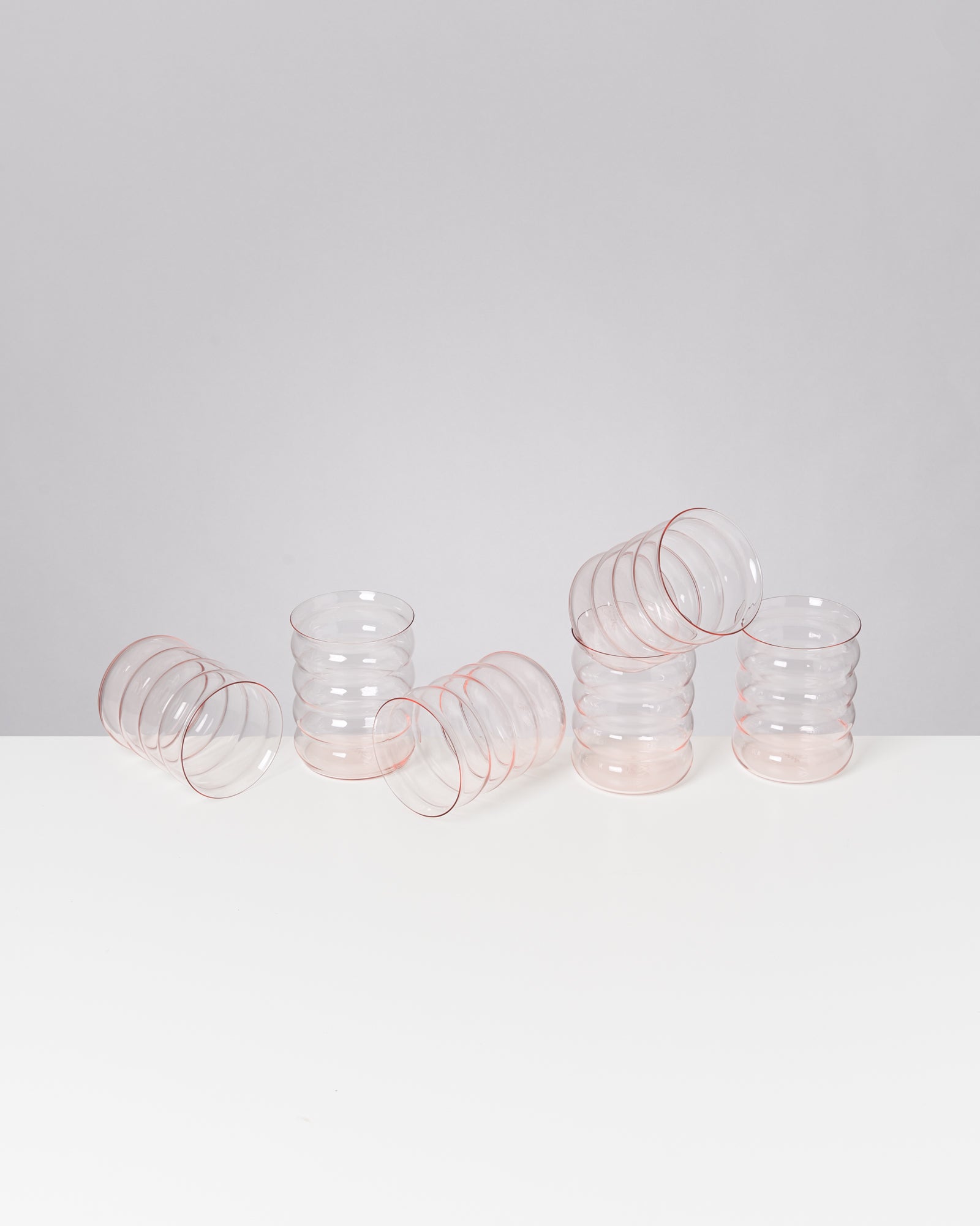 Sechs transparente Gläser mit rosa Rand stehen und liegen auf einem weißen Tisch vor grauem Hintergrund.