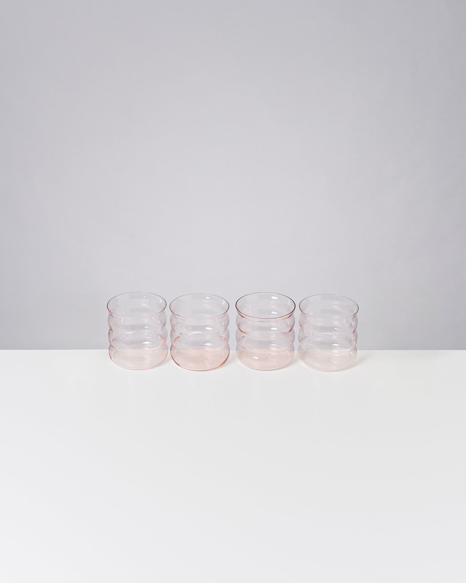 Vier transparent-rosafarbene Gläser stehen nebeneinander auf einer weißen Oberfläche vor grauem Hintergrund.