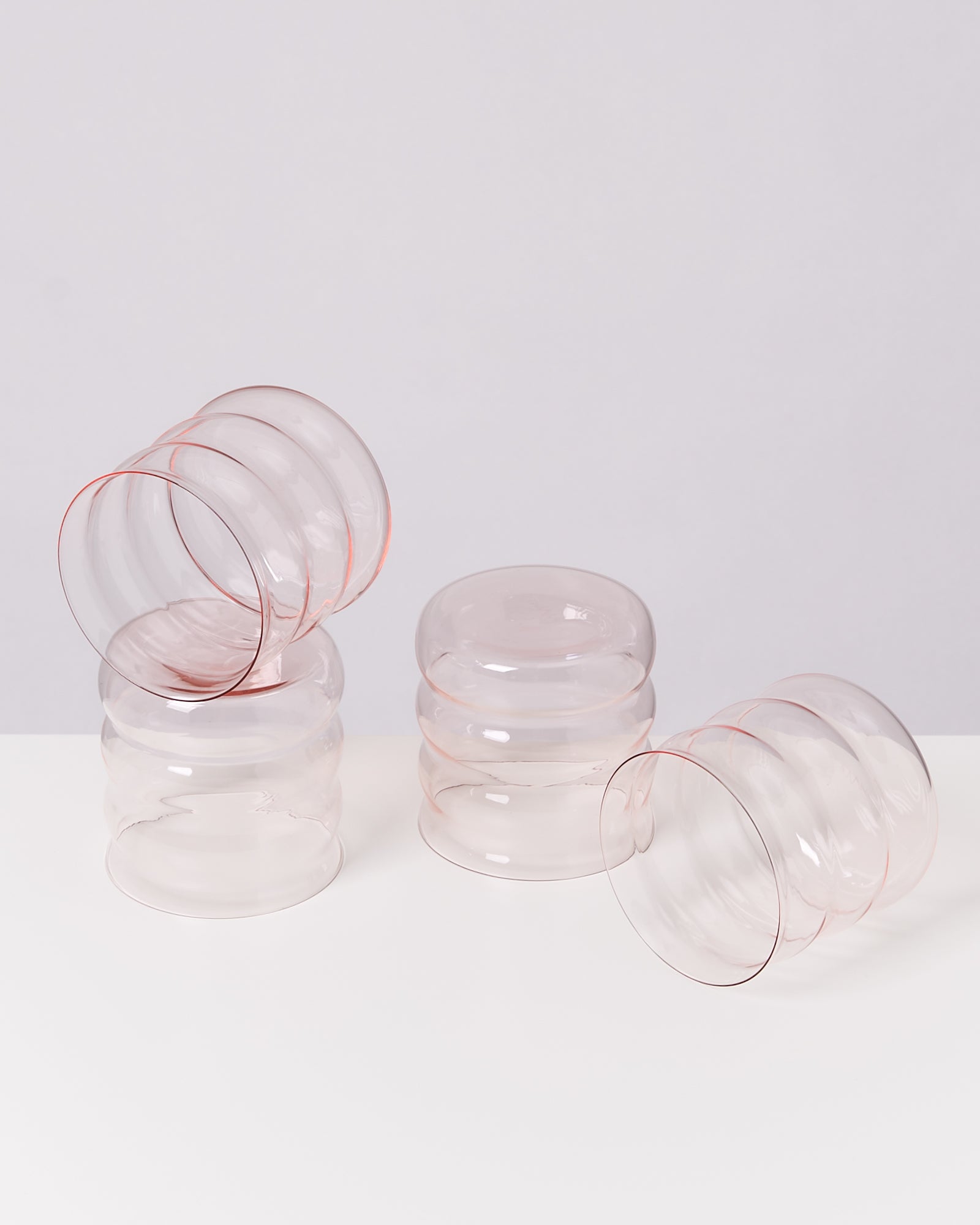Vier stapelbare, wellenförmige Glasbecher in transparentem Rosa auf einer weißen Oberfläche.