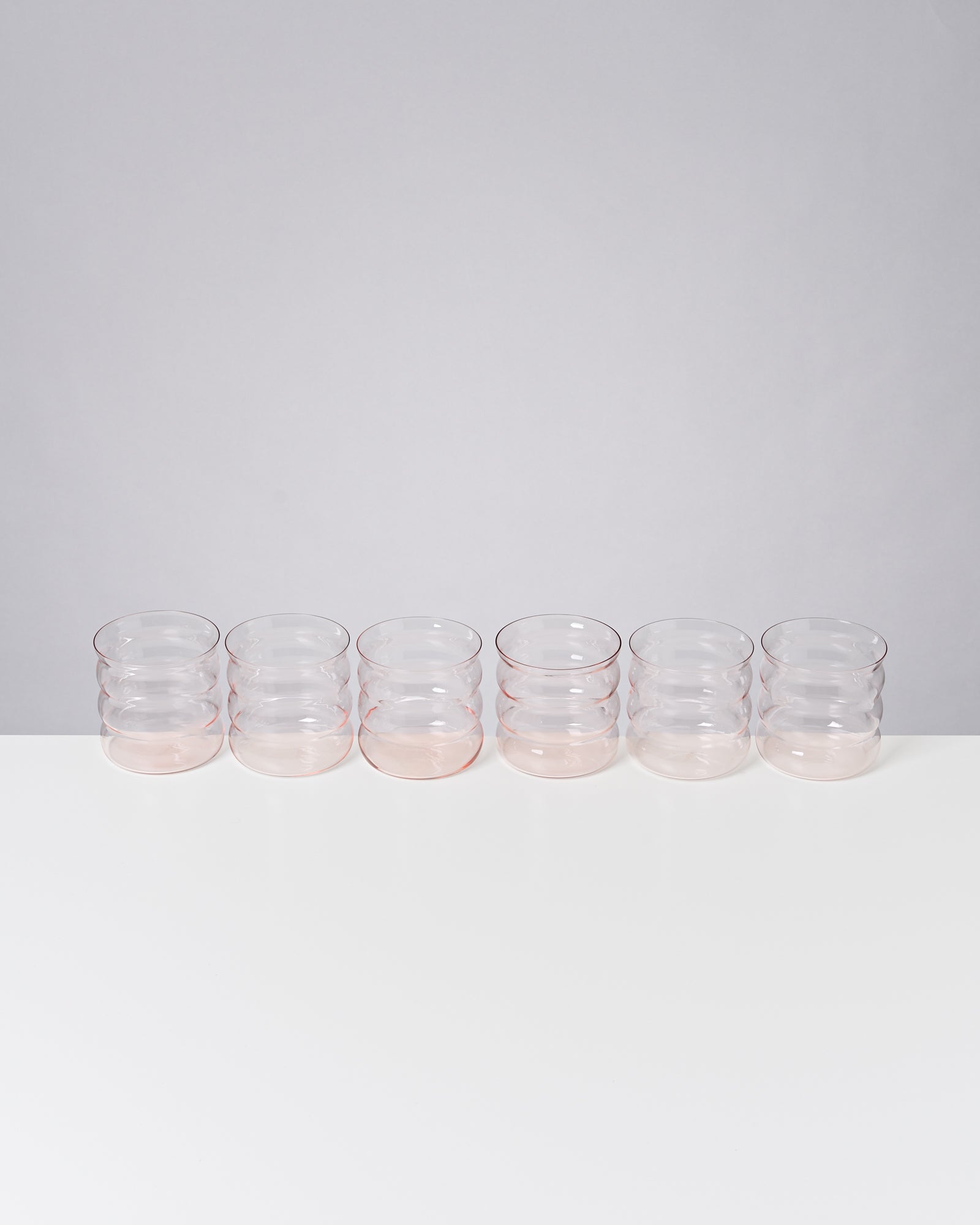 Sechs gestapelte, transparente Trinkgläser auf einem weißen Tisch vor einer grauen Wand.