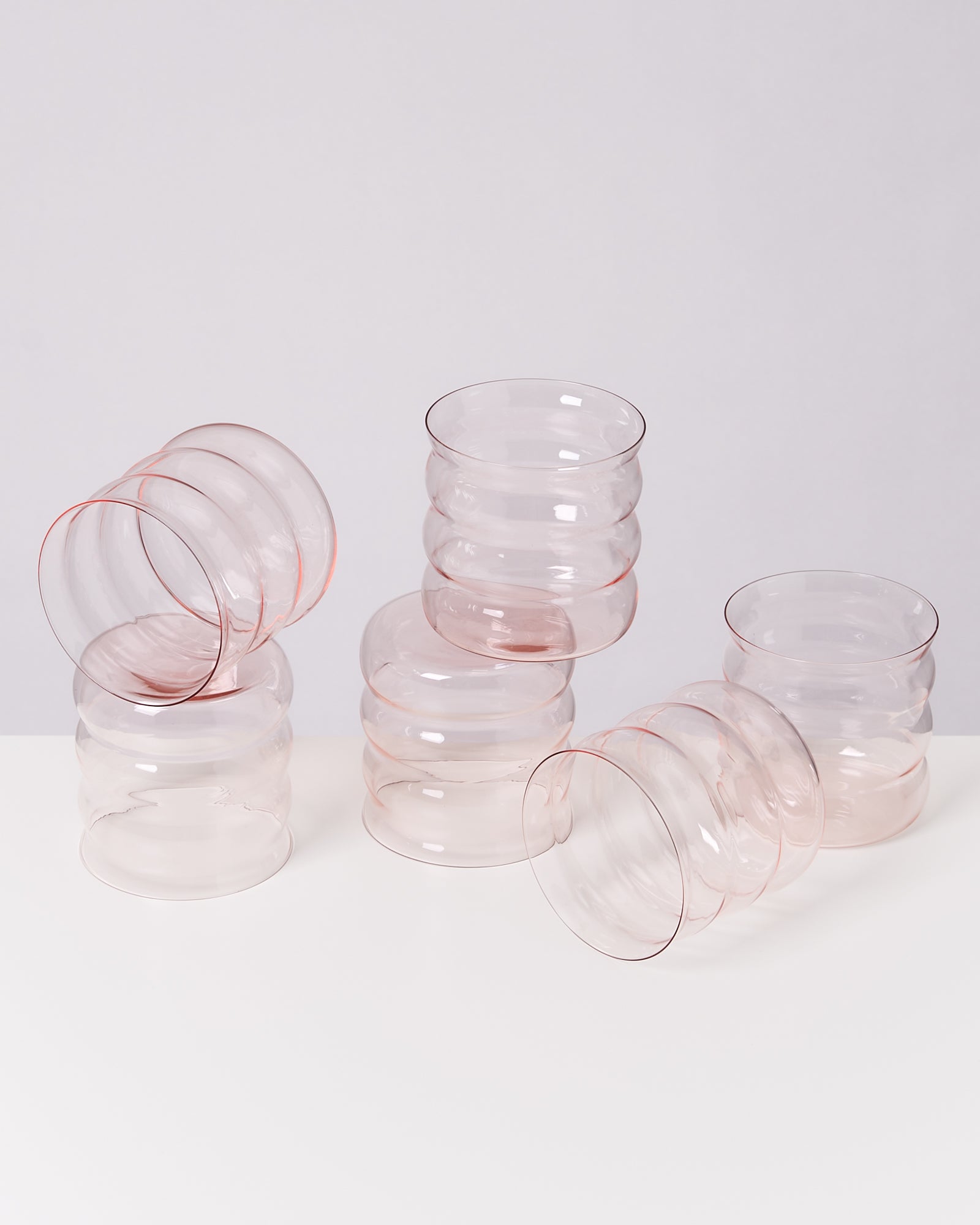 Sechs wellenförmige, durchsichtige Gläser mit leicht rosa Tönung auf einer weißen Fläche arrangiert.