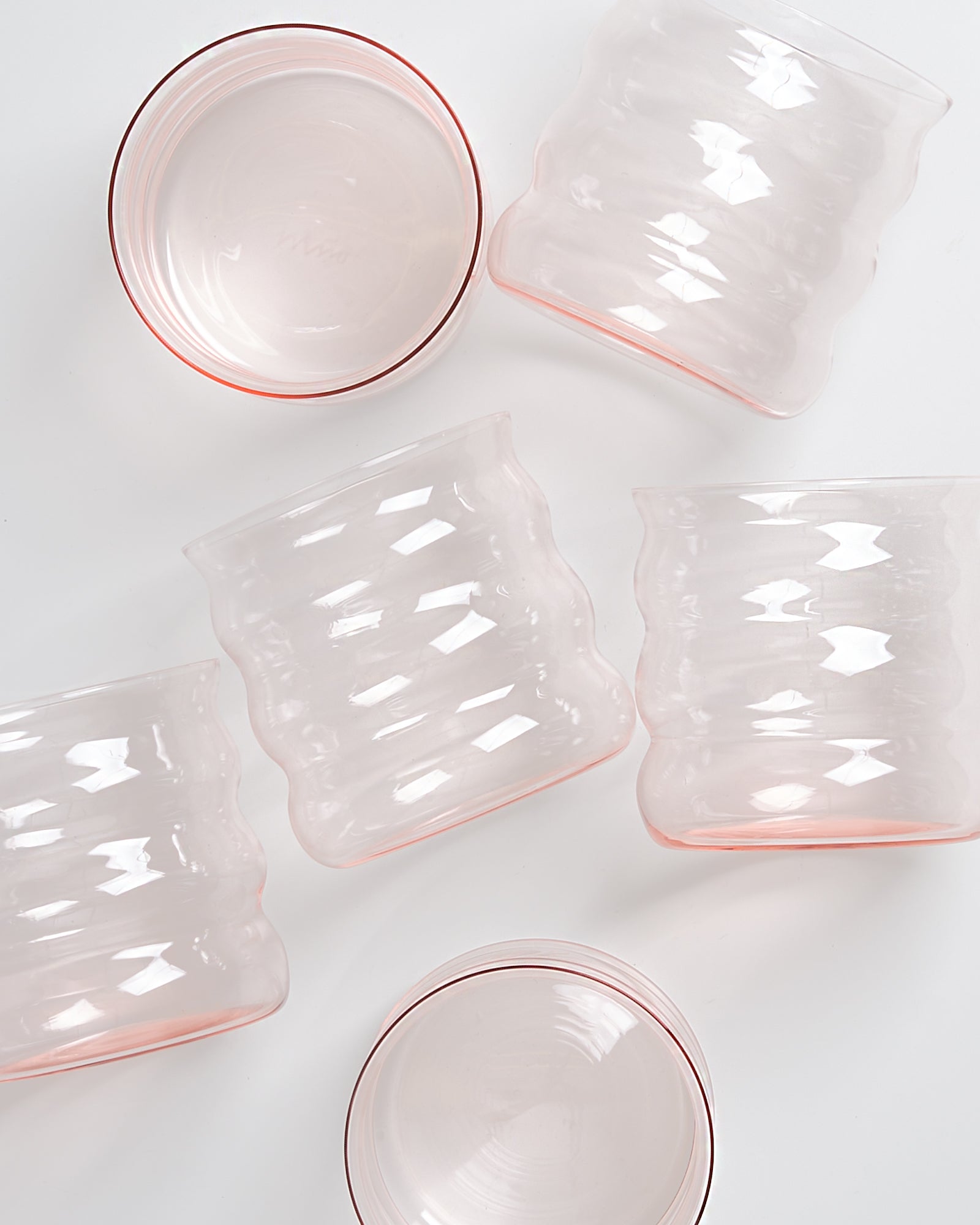 Fünf rosafarbene, gewellte Glasbecher liegen unregelmäßig auf einer weißen Oberfläche verteilt.