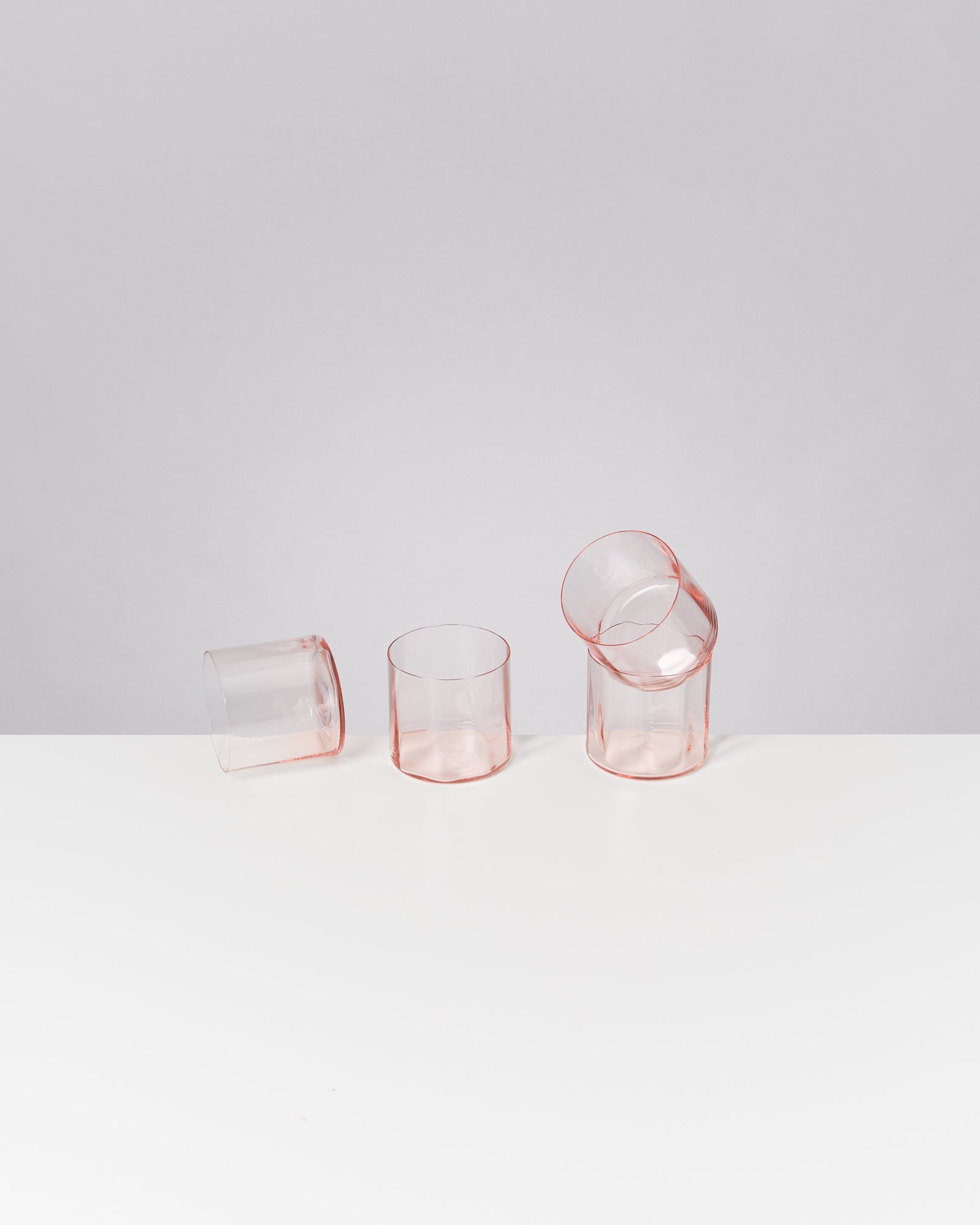 Drei transparente rosa Gläser, zwei stehend und eines umgekippt, auf einem weißen Tisch vor grauem Hintergrund.