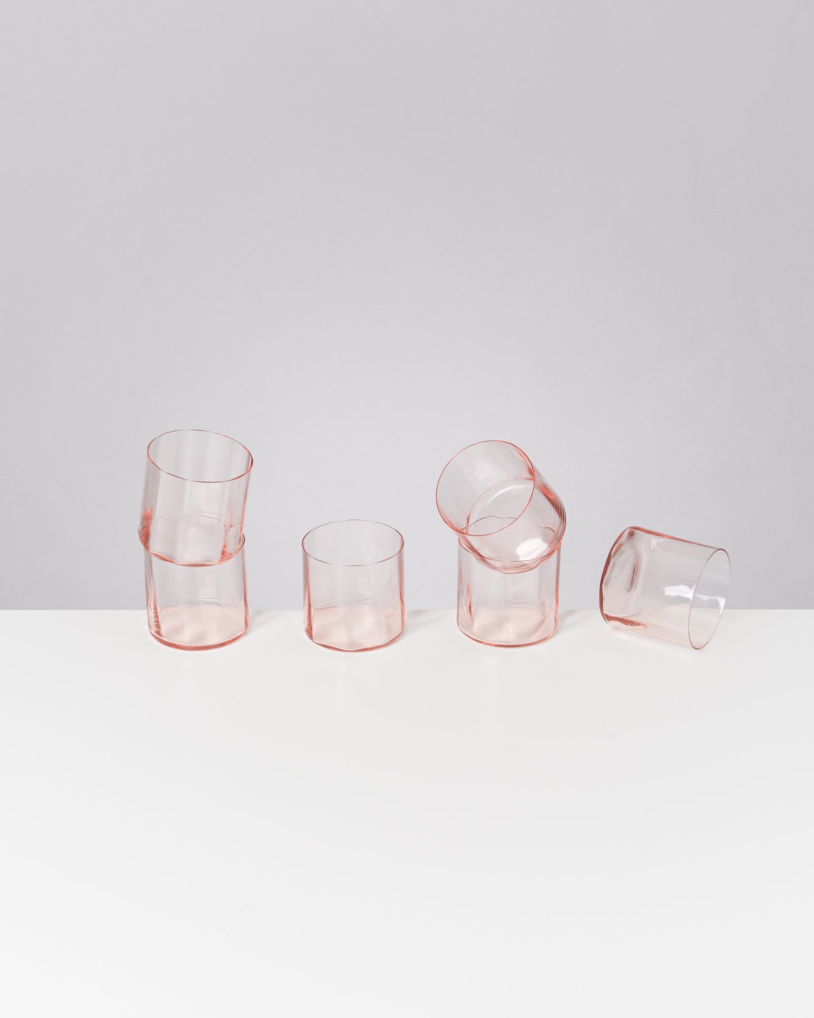 Fünf rosafarbene Gläser stehen in einer unregelmäßigen Formation auf einer weißen Oberfläche vor grauem Hintergrund.