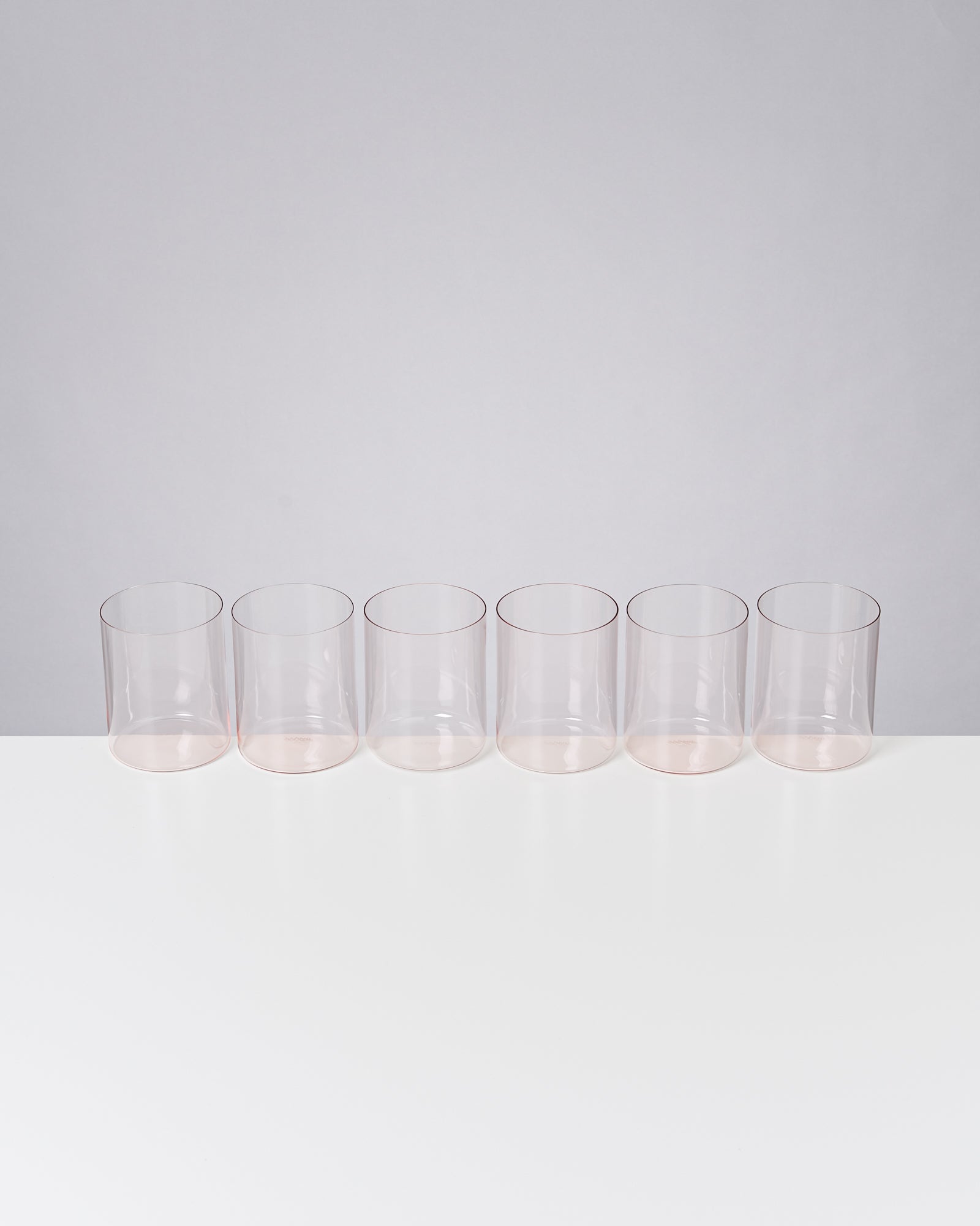 Sechs leere, durchsichtige Gläser stehen in einer Reihe auf einer weißen Oberfläche vor einem grauen Hintergrund.