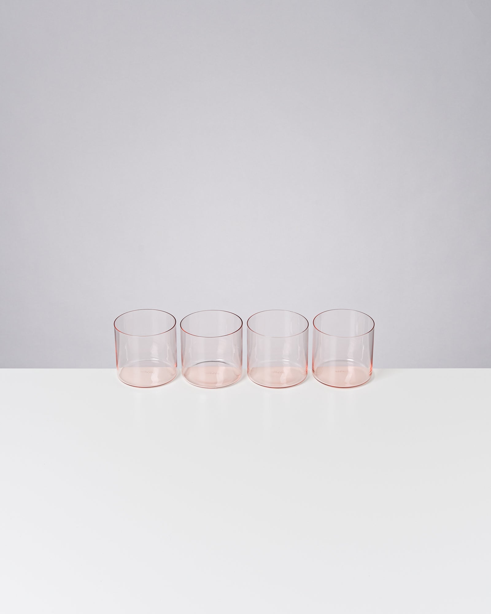 Vier transparente, rosafarbene Gläser stehen in einer Reihe auf einem weißen Tisch vor grauem Hintergrund.