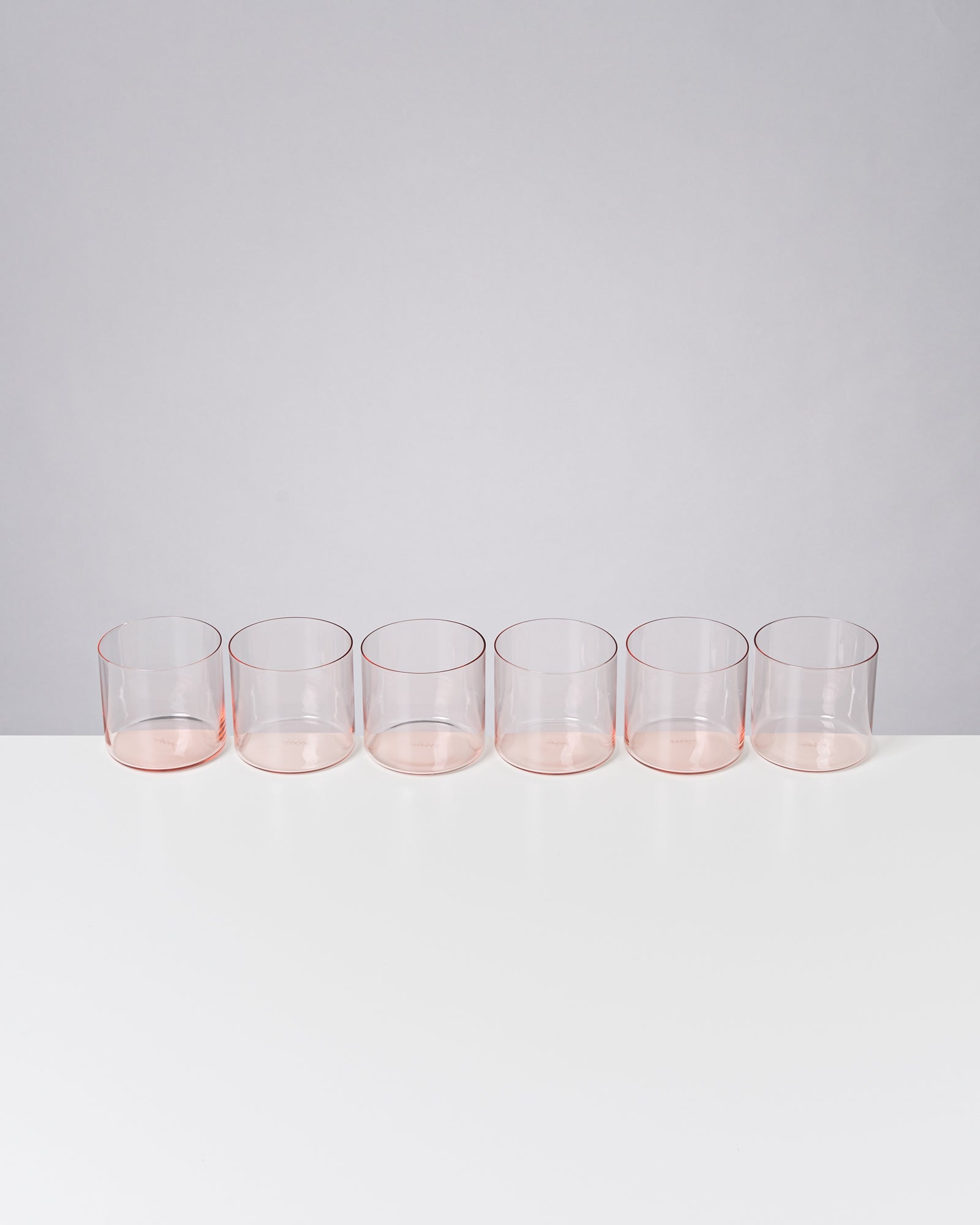 Sechs leere, transparente Trinkgläser in einer Reihe auf weißem Untergrund vor grauem Hintergrund.