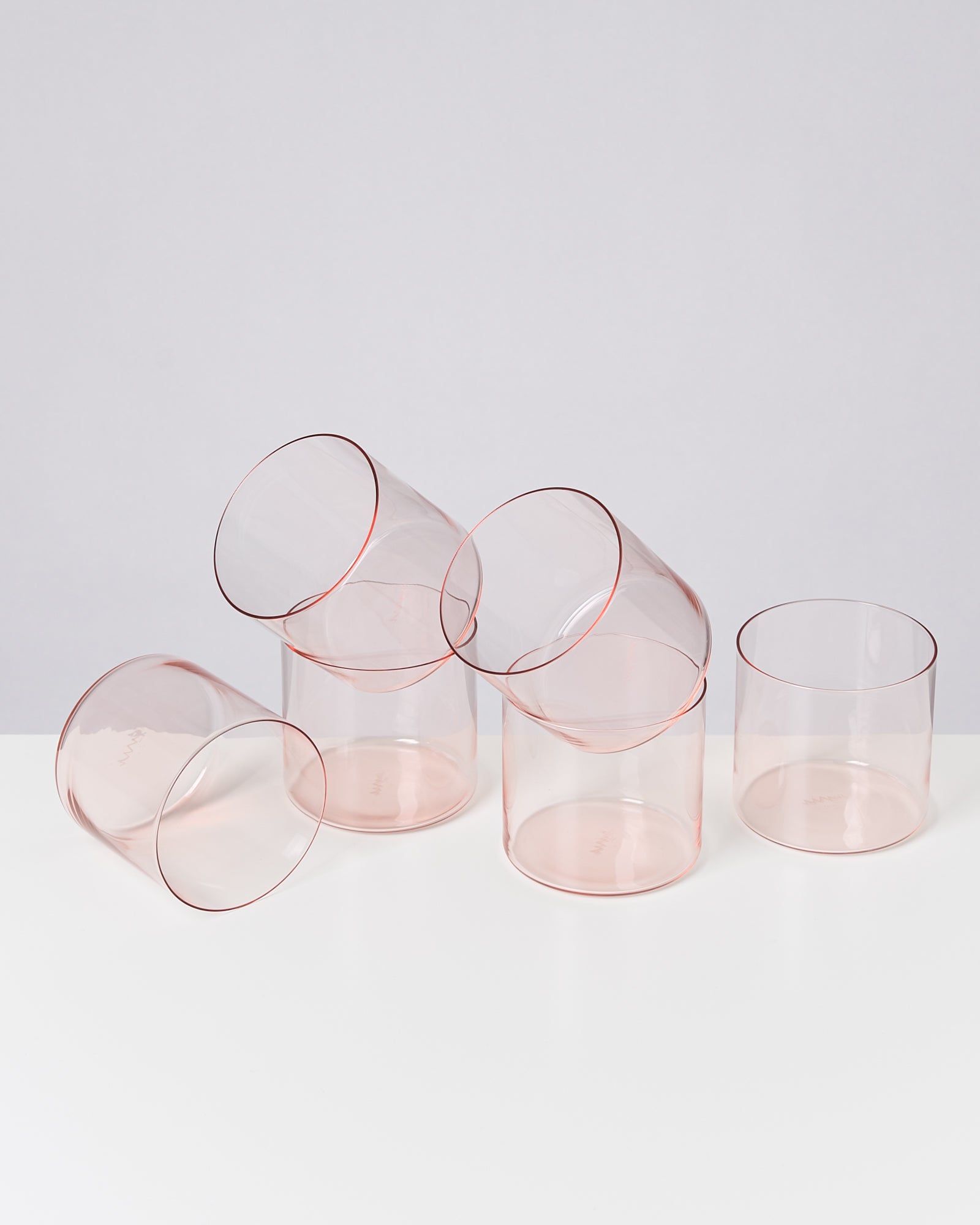 Fünf durchsichtige, rosa Tumbler-Gläser stehen und liegen auf einem weißen Tisch vor einem grauen Hintergrund.