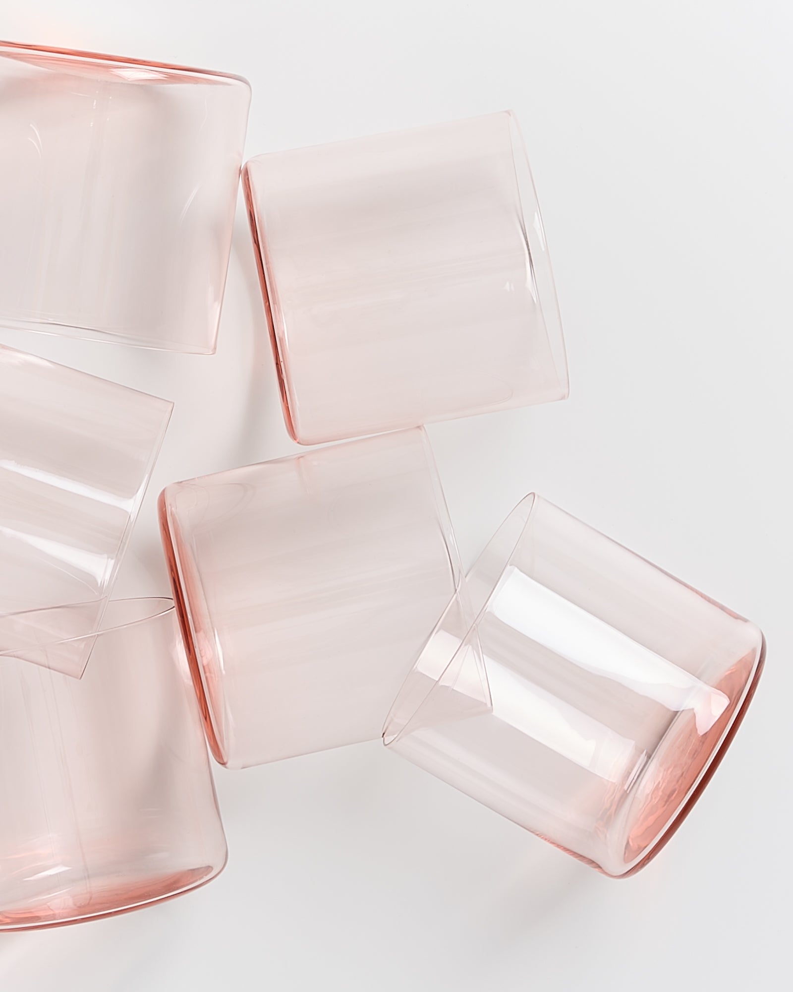 Sechs rosafarbene, transparente Trinkgläser liegen schräg auf einer weißen Oberfläche.