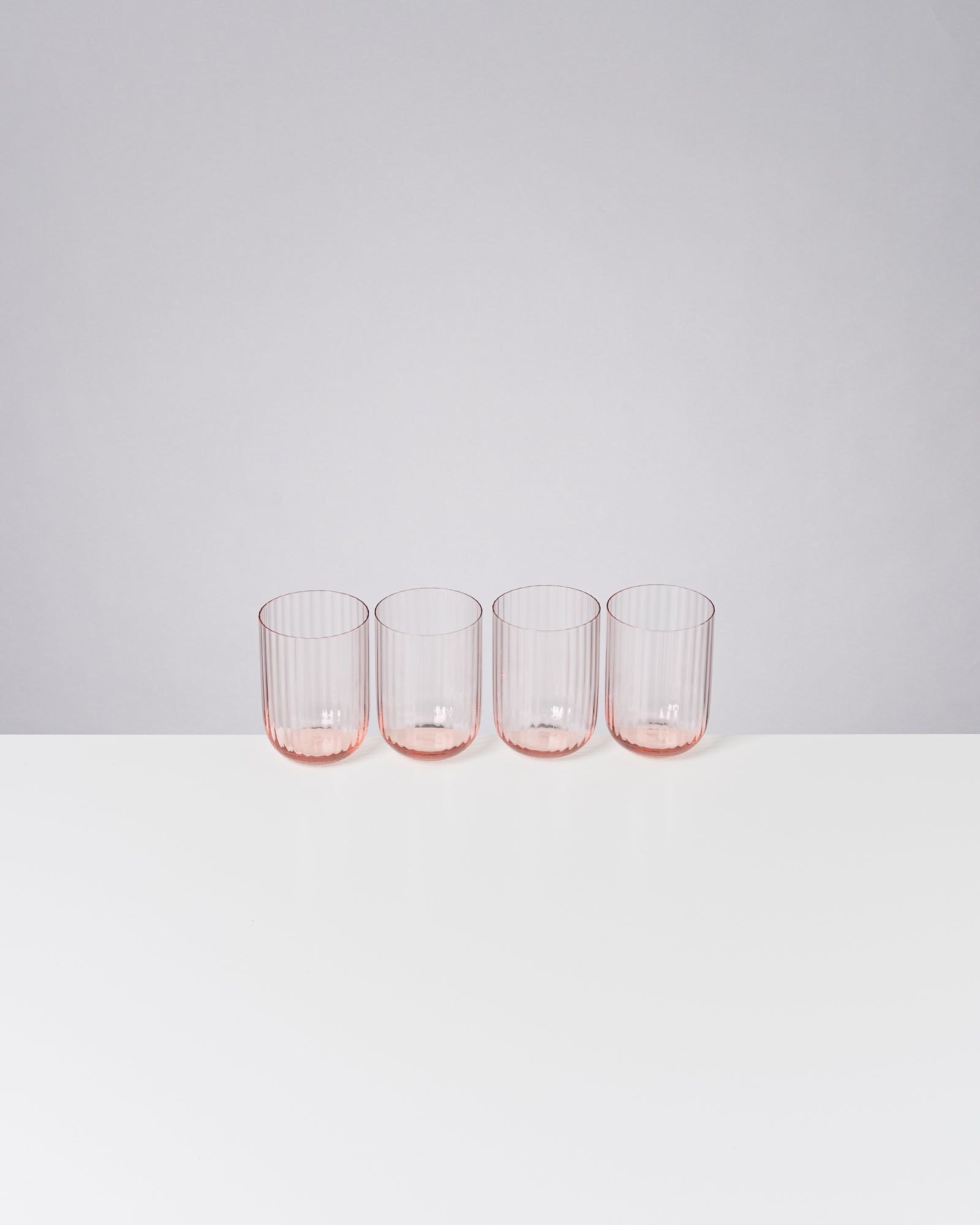 Vier gerippte, rosa Gläser stehen in einer Reihe auf einem weißen Untergrund vor grauem Hintergrund.